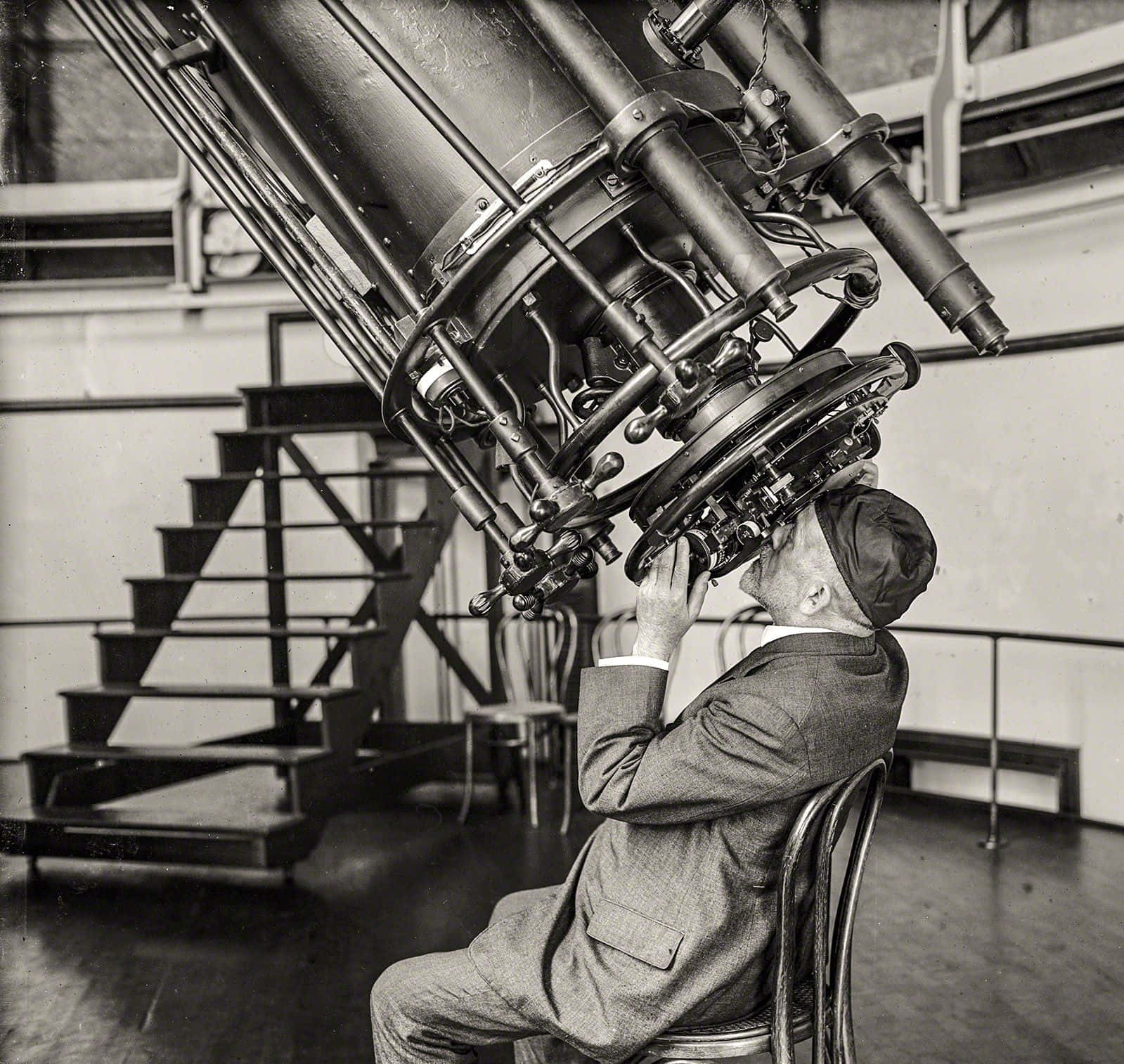 astronomy 桌布 壁紙 — 一名身穿西裝的男子在光線昏暗的天文台透過一個巨大的復古望遠鏡觀看，背景有樓梯。