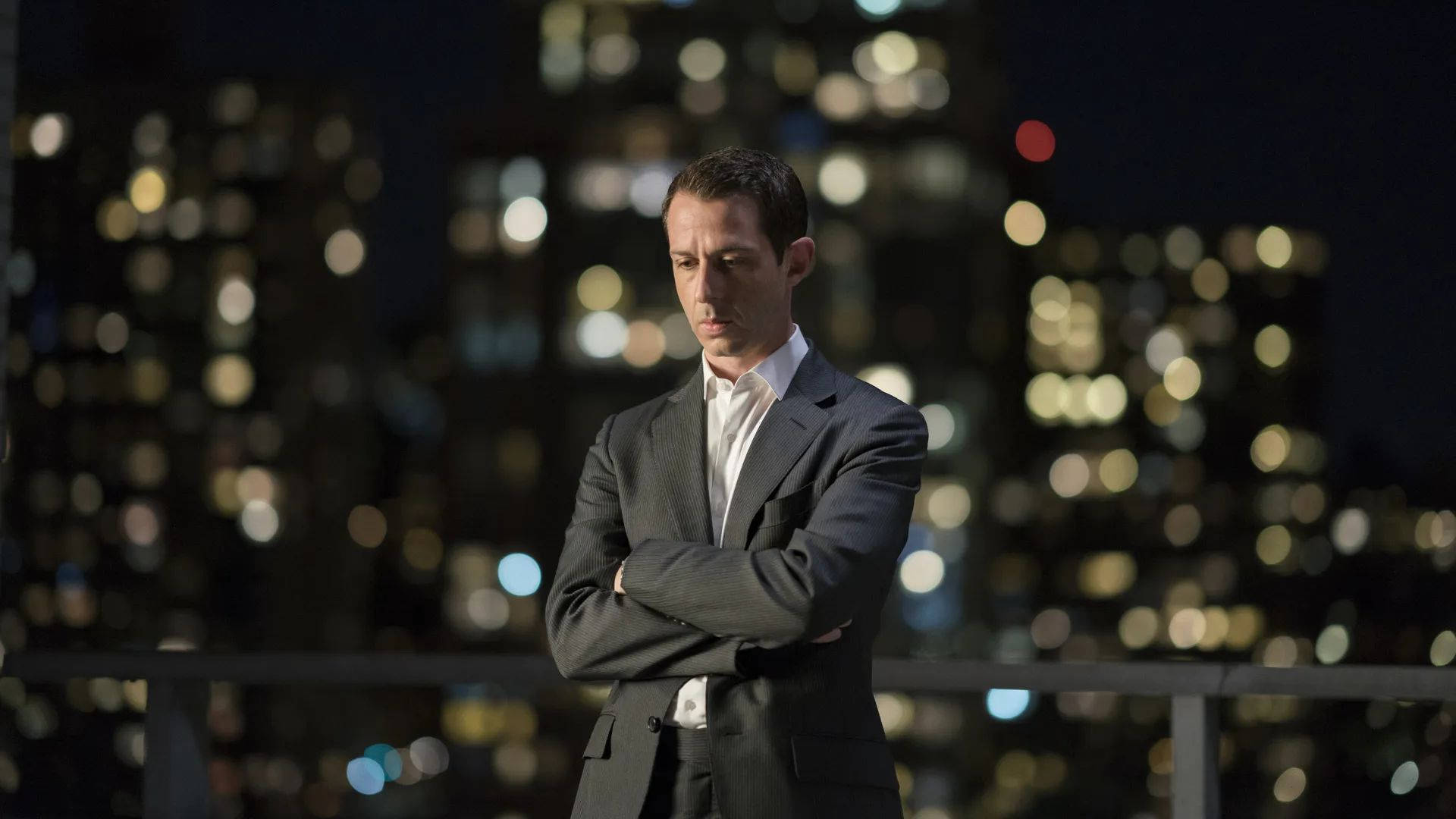Succession 桌布 壁紙 — 一名身穿西裝的男子在夜晚的屋頂上交叉雙臂站立，俯視著模糊的都市天際線與閃爍燈光。
