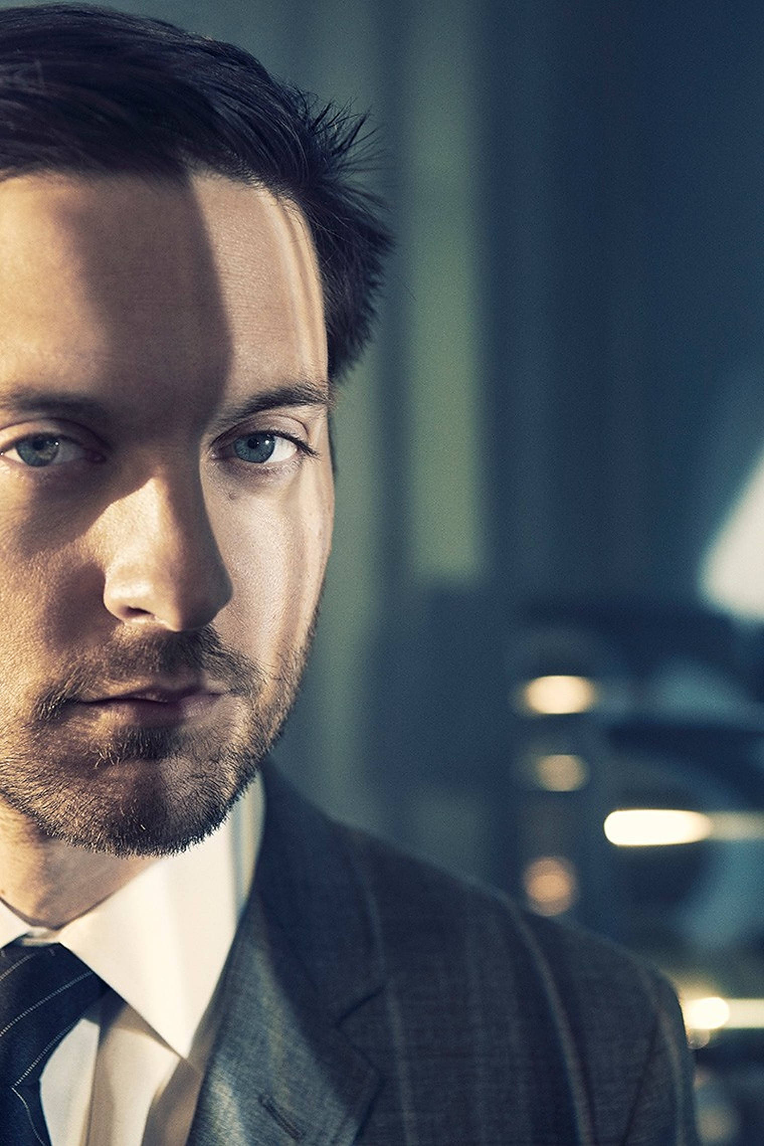 Tobey Maguire 桌布 壁紙 — 一名身穿西裝的男子在戲劇性燈光下，以模糊的辦公室背景為襯托，專注地凝視鏡頭。