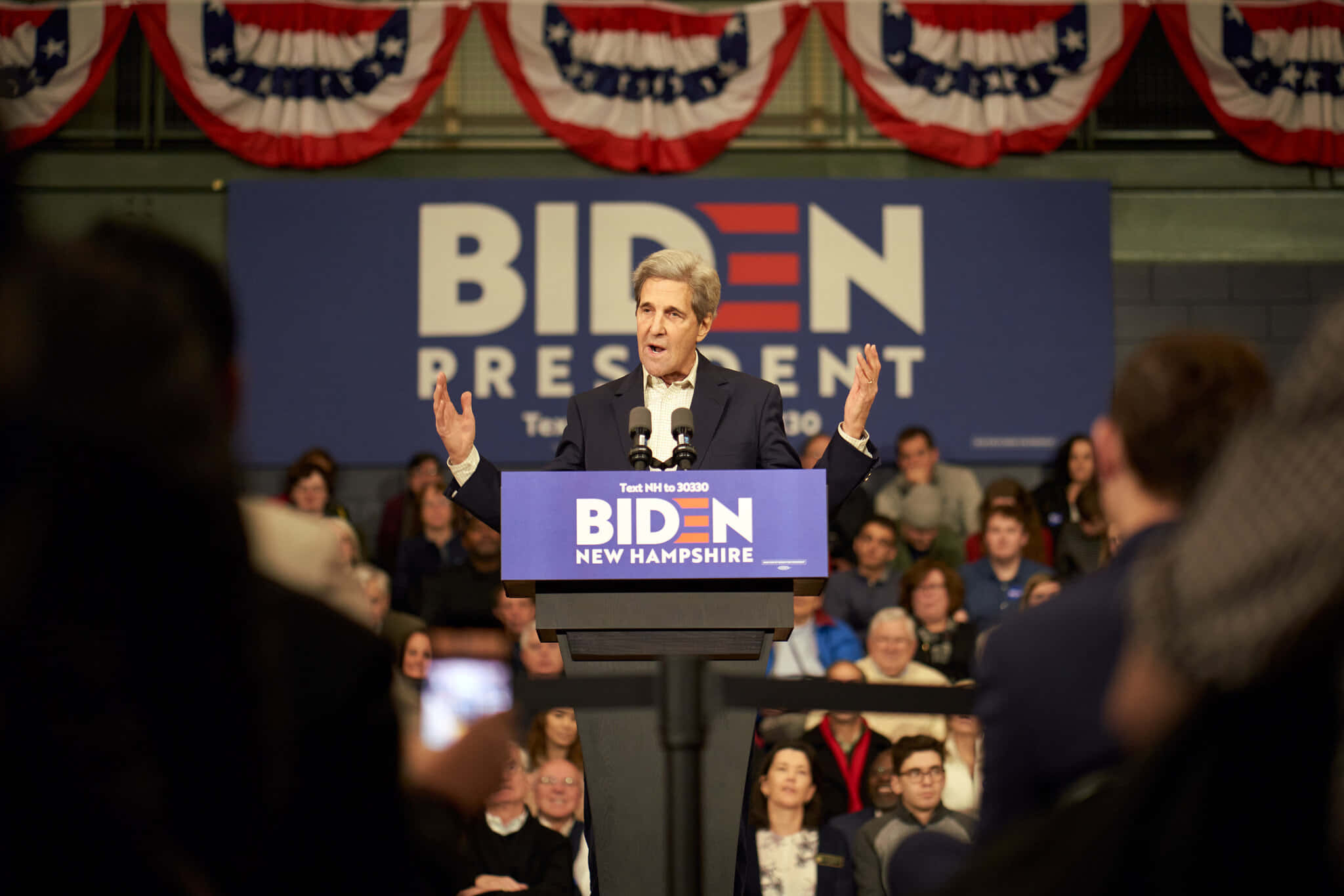 喬·拜登 桌布 壁紙 — 一名身穿西裝的男子在拜登總統競選講台上熱情演講，頭頂懸掛著美國國旗裝飾。