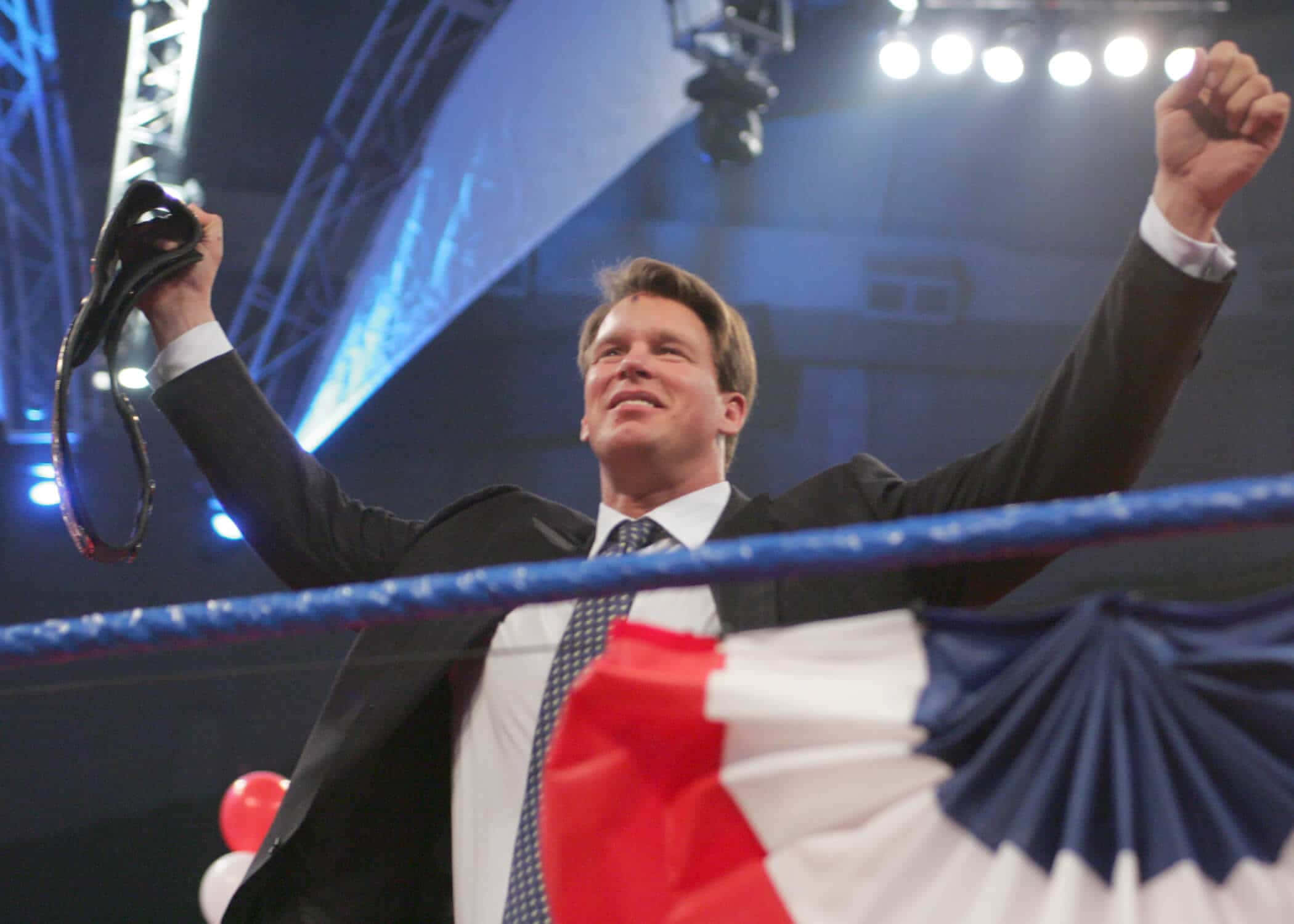 WWE 桌布 壁紙 — 一名身穿西裝的男子在明亮的舞台燈光下，於摔角擂台內勝利地高舉拳頭和冠軍腰帶。