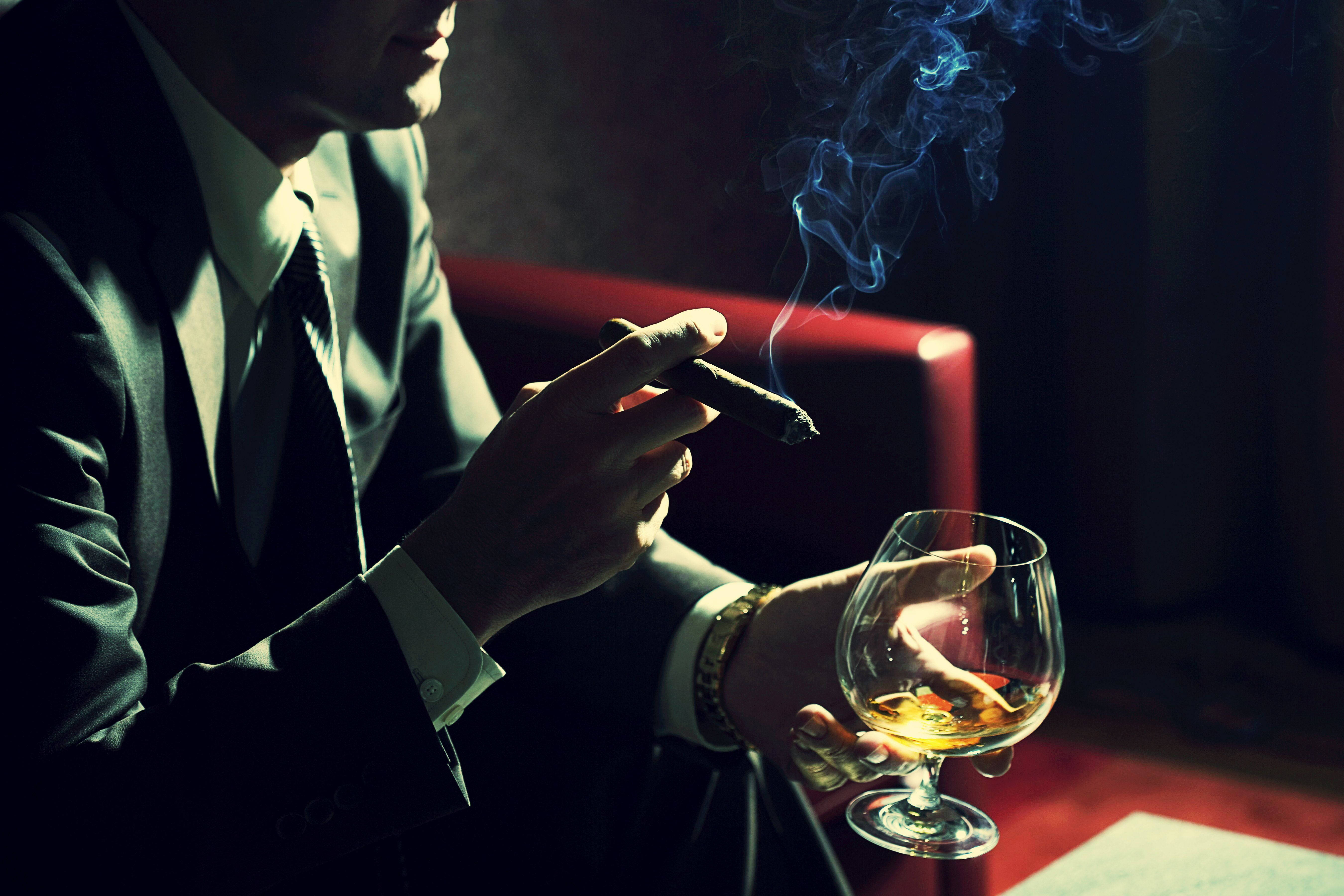 Drink 桌布 壁紙 — 一名身穿西裝的男子在昏暗的房間裡抽雪茄，手持一杯威士忌，藍色煙霧繚繞。