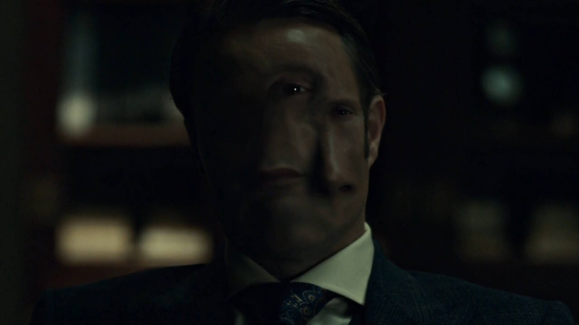 Mads Mikkelsen 桌布 壁紙 — 一名身穿西裝的男子在昏暗的房間裡專注地凝視，身後是模糊的書架。
