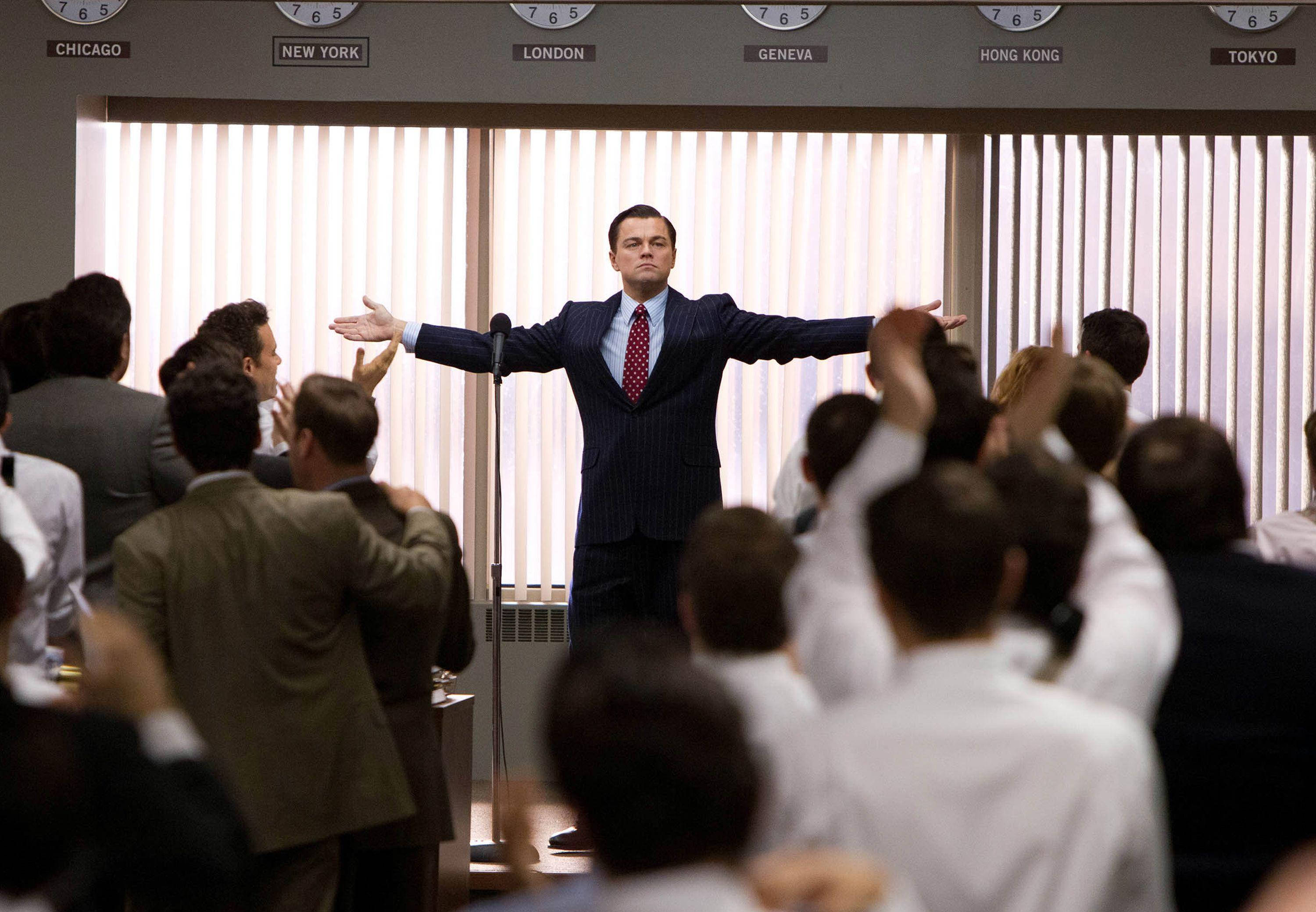 Wolf Of Wall Street 桌布 壁紙 — 一名身穿西裝的男子在會議室中，於全球城市時鐘下方，張開雙臂站在人群前。