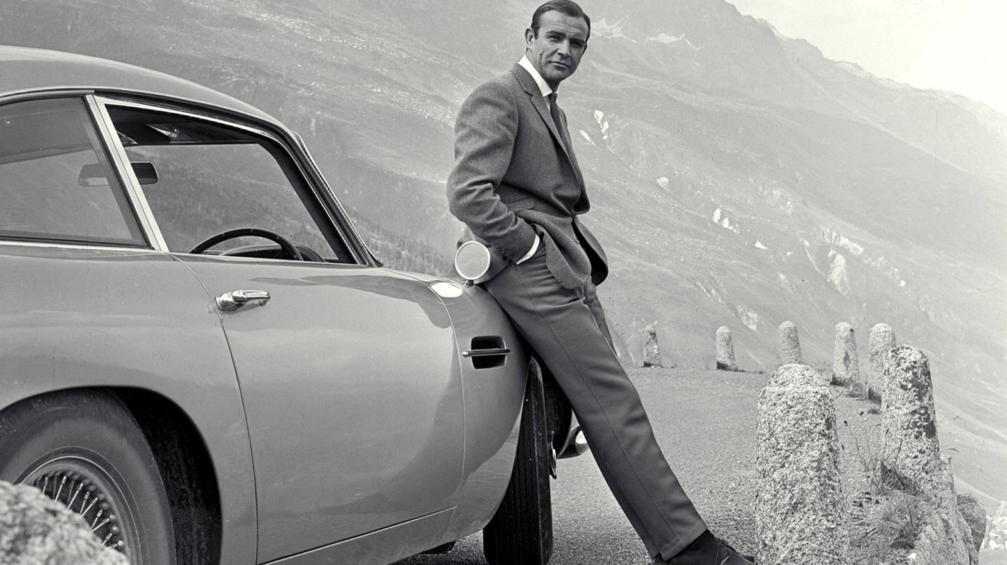 Sean Connery 桌布 壁紙 — 一名身穿西裝的男子在有一道石欄杆和遠處山峰的山路上靠著一輛復古跑車。