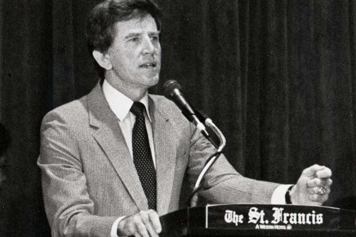 桌布 壁紙 — 一名身穿西裝的男子在標有 The St Francis hotel 的講台上，對著麥克風充滿熱情地演講。
