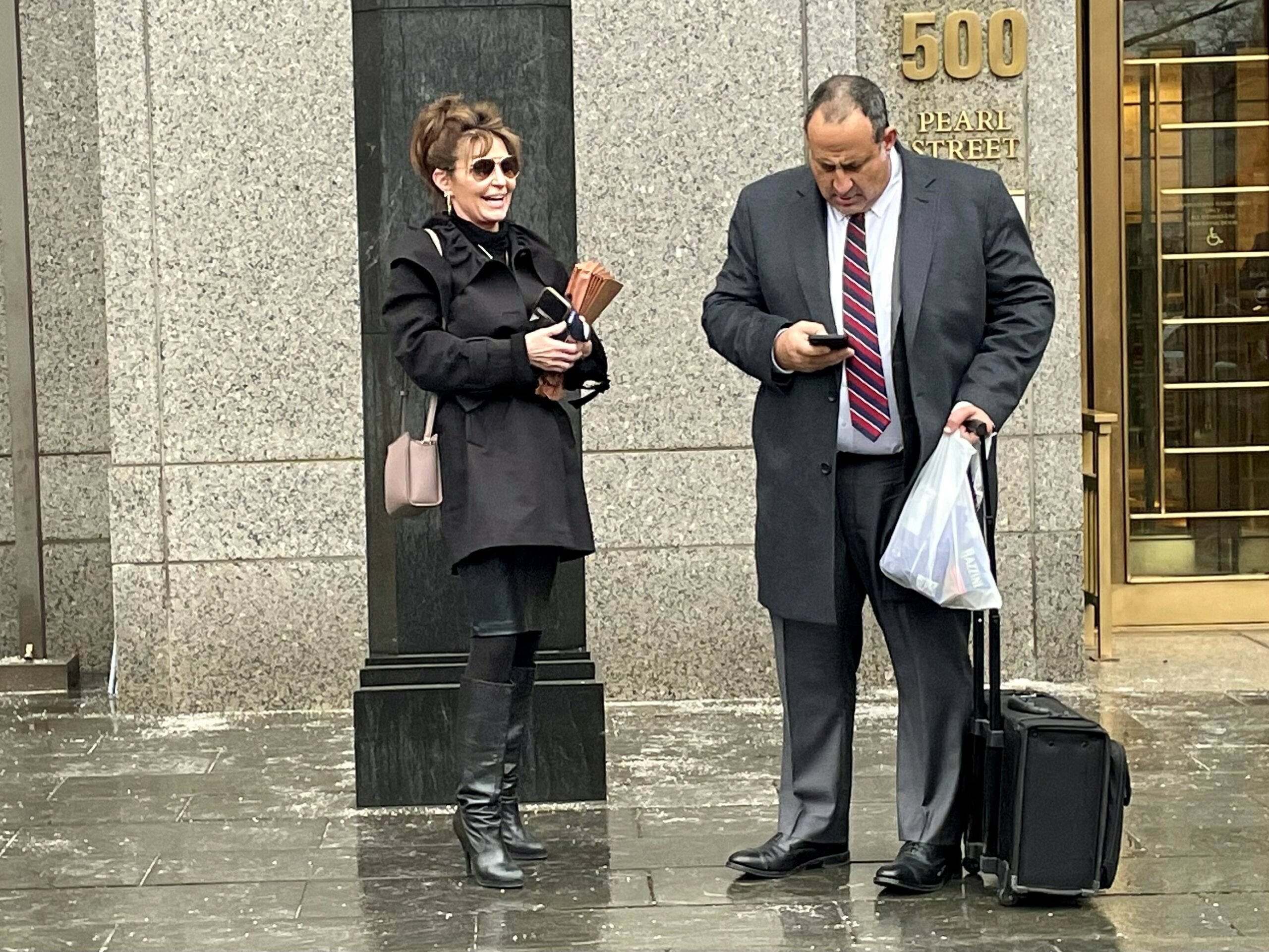 Lawyer 桌布 壁紙 — 一名身穿西裝的男子在標有500 Pearl Street的建築物外，拖著行李箱查看手機，附近站著一位身穿黑色外套和太陽眼鏡的女子。