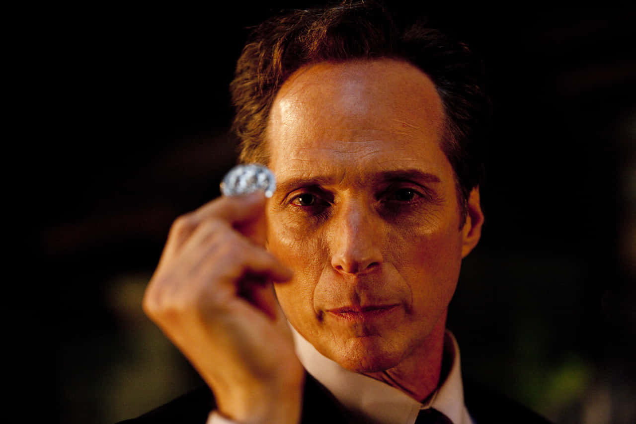 William Fichtner 桌布 壁紙 — 一名身穿西裝的男子在深色背景下將一顆閃閃發光的鑽石舉到眼前。