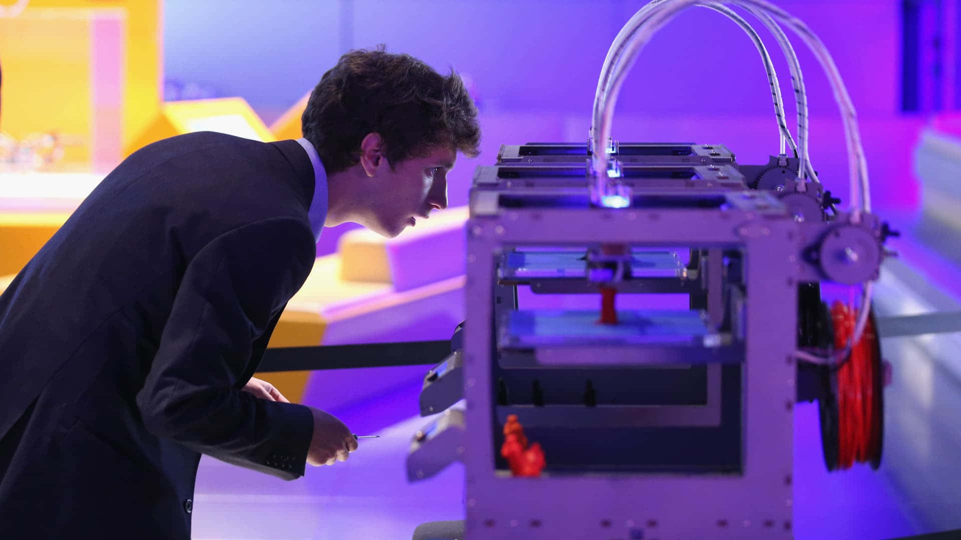 3d Printer 桌布 壁紙 — 一名身穿西裝的男子在現代科技展覽中，專注地俯身靠近一部散發著紫色和藍色光芒的3D列印機。