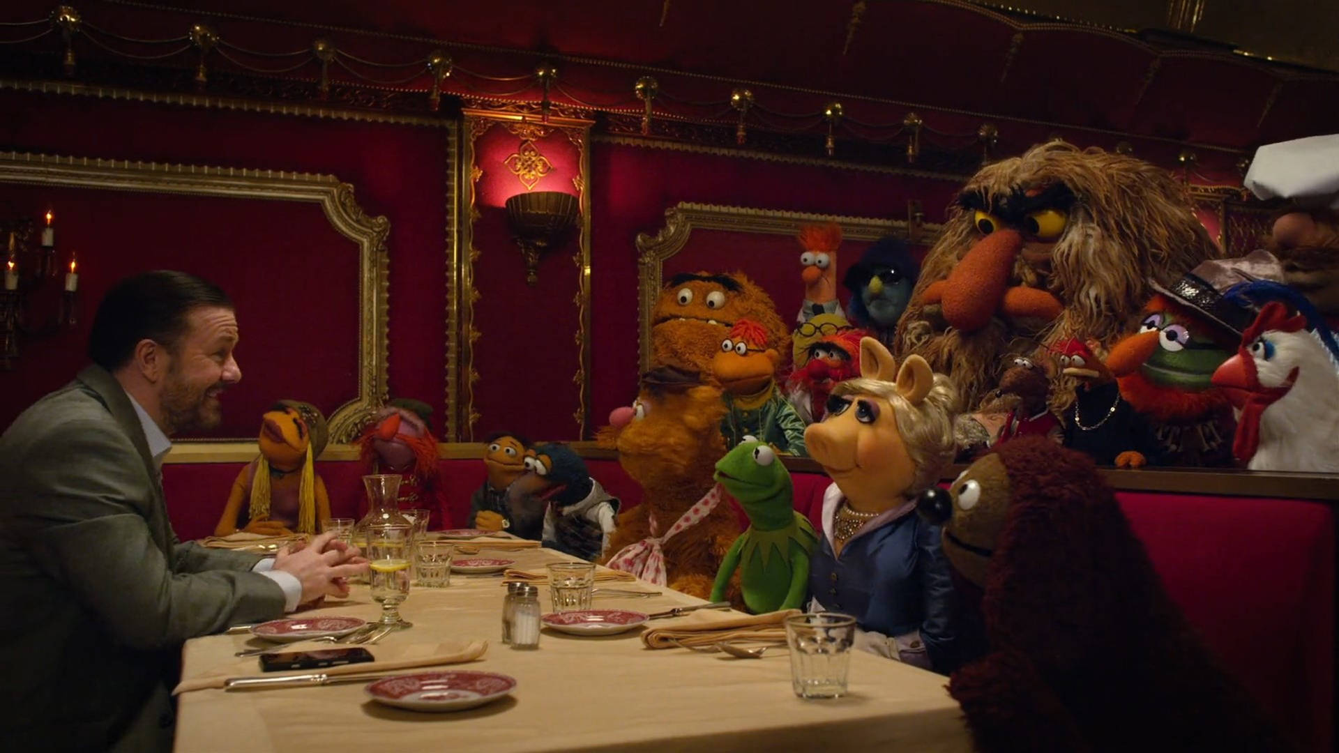 muppets 桌布 壁紙 — 一名身穿西裝的男子在裝飾華麗的紅色餐廳卡座中，被色彩繽紛的Muppets包圍，享用豐盛的晚餐。