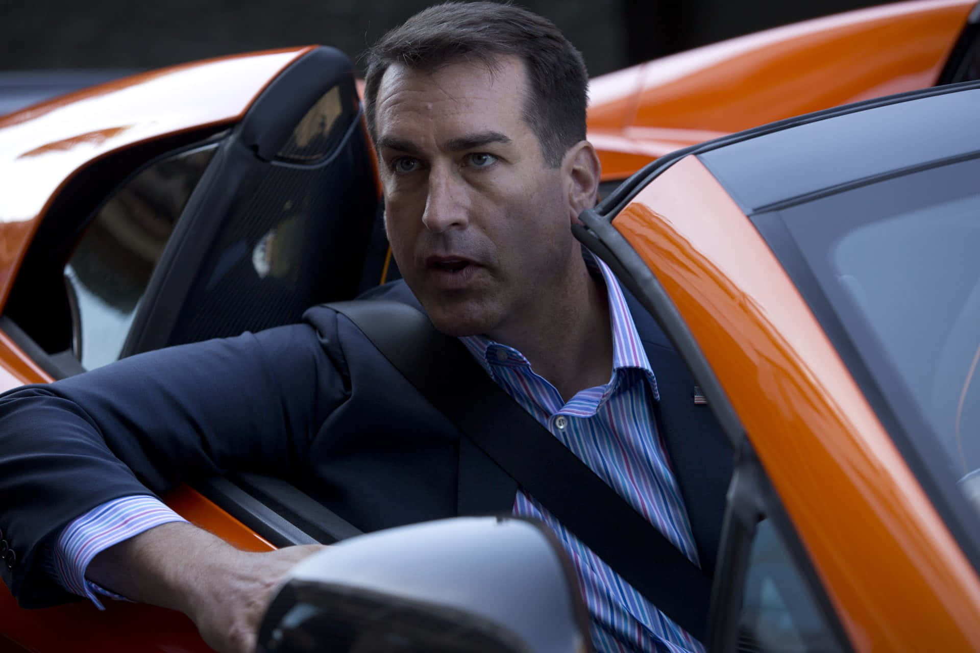 Rob Riggle 桌布 壁紙 — 一名身穿西裝的男子坐在亮橙色跑車內，表情驚訝。