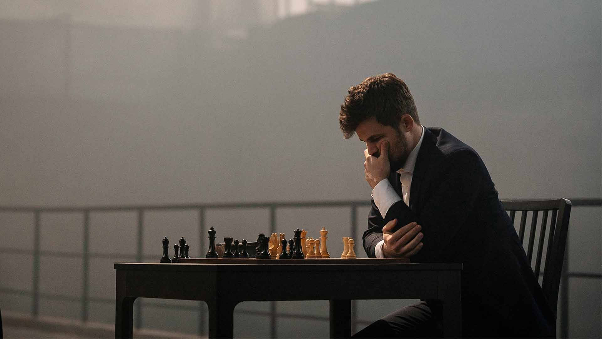 Magnus 桌布 壁紙 — 一名身穿西裝的男子坐在棋盤桌前，手托下巴陷入沉思，陽光透過他身後朦朧的窗戶灑進來。