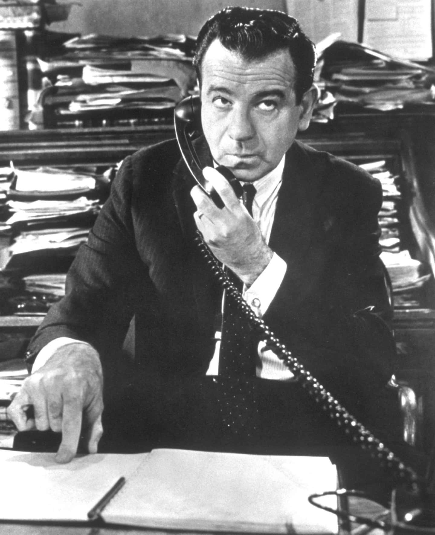 Walter Matthau 桌布 壁紙 — 一名身穿西裝的男子坐在雜亂的辦公桌前，手持電話聽筒，表情嚴肅。