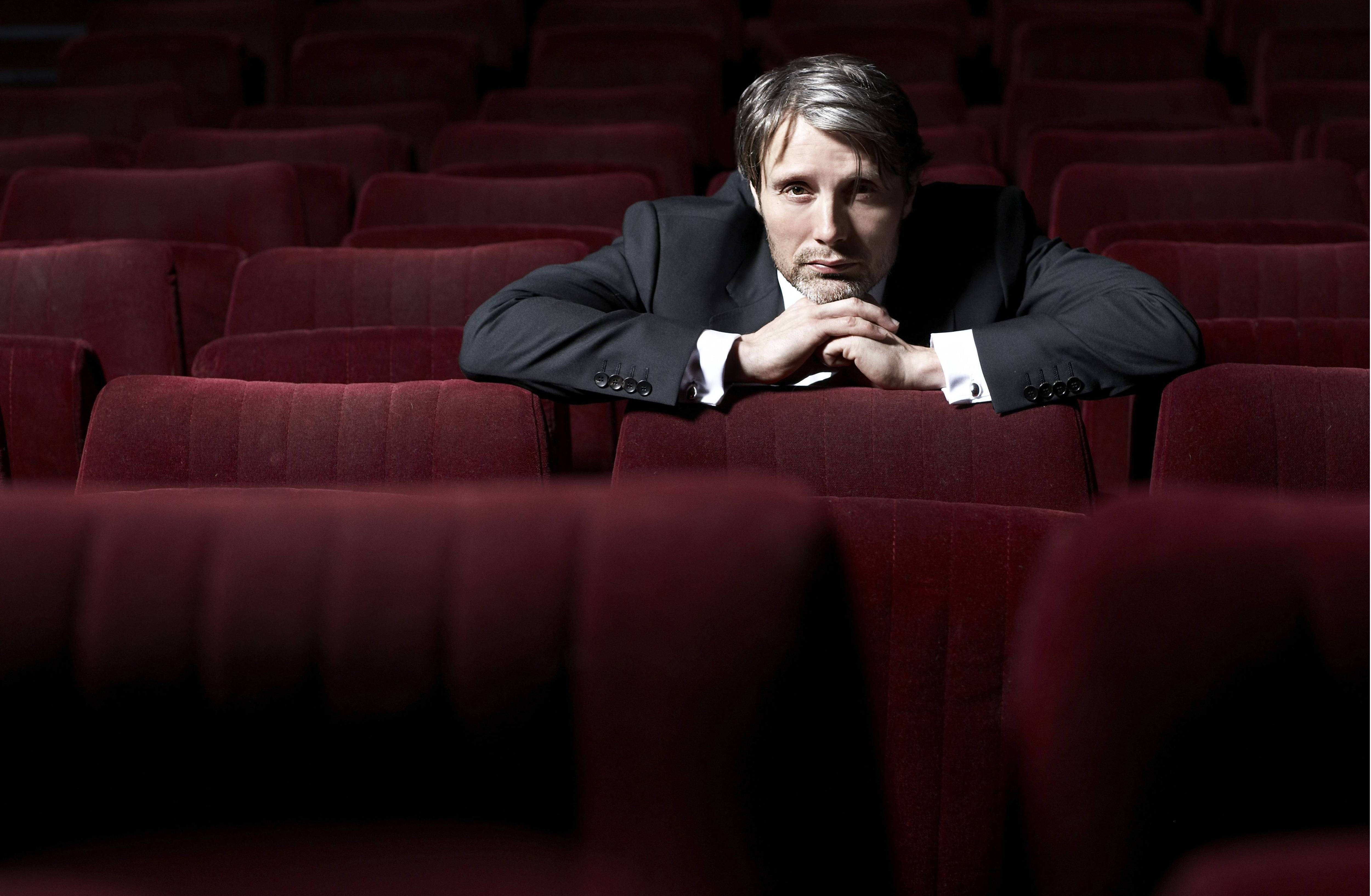 Mads Mikkelsen 桌布 壁紙 — 一名身穿西裝的男子將下巴擱在交疊的雙手上，放在紅色劇院座位上，在空蕩的禮堂中若有所思地凝視鏡頭。