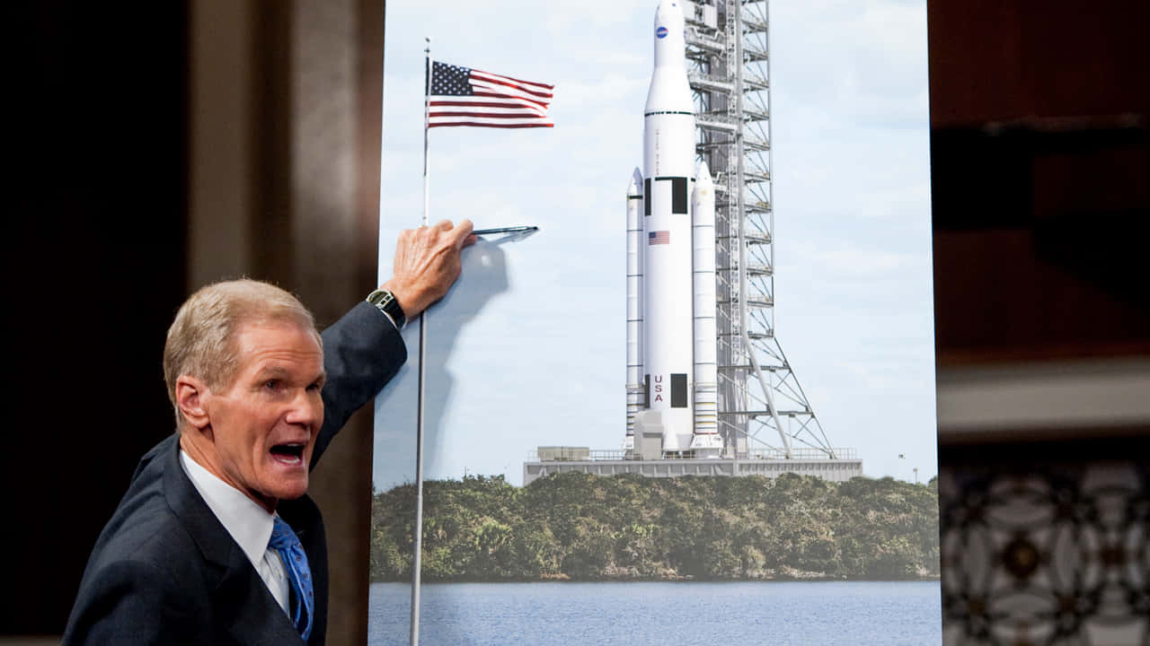 NASA 動態圖片 桌布 壁紙 — 一名身穿西裝的男子戲劇性地向一張大型海報做手勢，海報上是美國太空總署的火箭在發射台上，美國國旗飄揚。