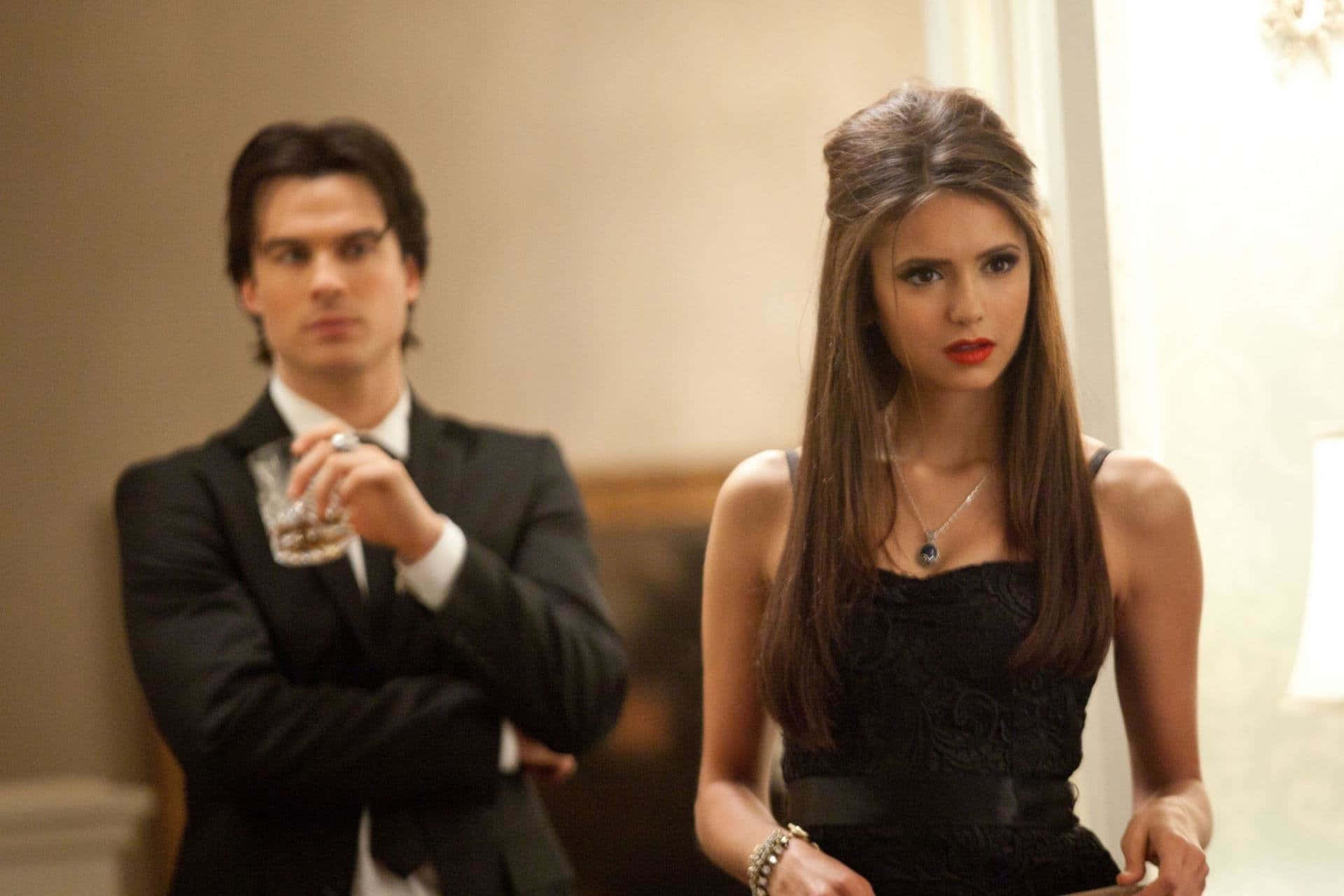 Vampire Diaries Desktop 桌布 壁紙 — 一名身穿西裝的男子手持酒杯，而一名身穿黑色連衣裙的女子則站在附近，身處優雅的室內環境。