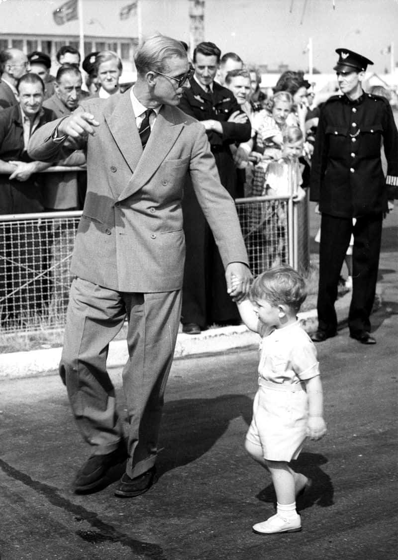 王子 桌布 壁紙 — 一名身穿西裝的男子牽著一個小孩的手，穿過戶外活動的人群。