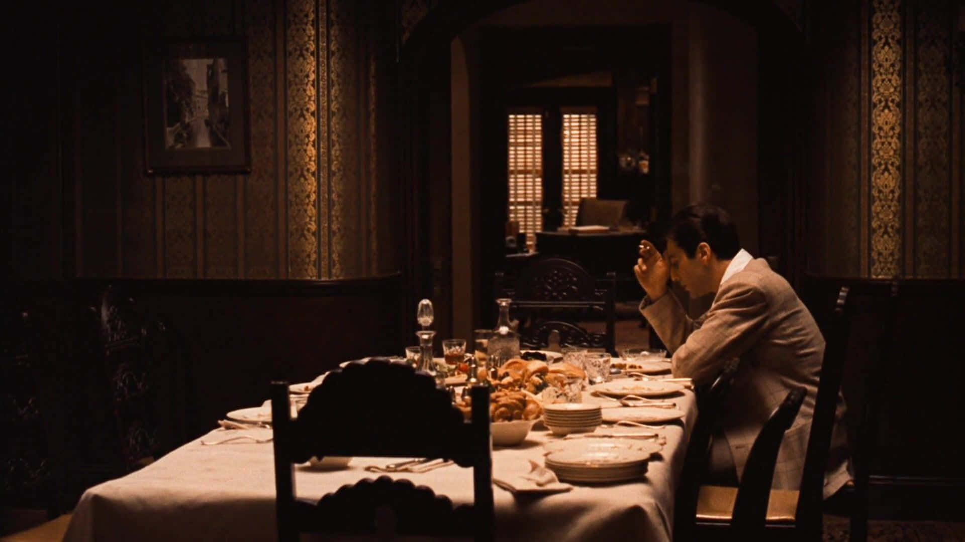 The Godfather 桌布 壁紙 — 一名身穿西裝的男子獨自坐在燈光昏暗、裝飾華麗的房間裡一張佈置奢華的餐桌旁，雙手抱頭，周圍是未動過的食物和溫暖的環境光。