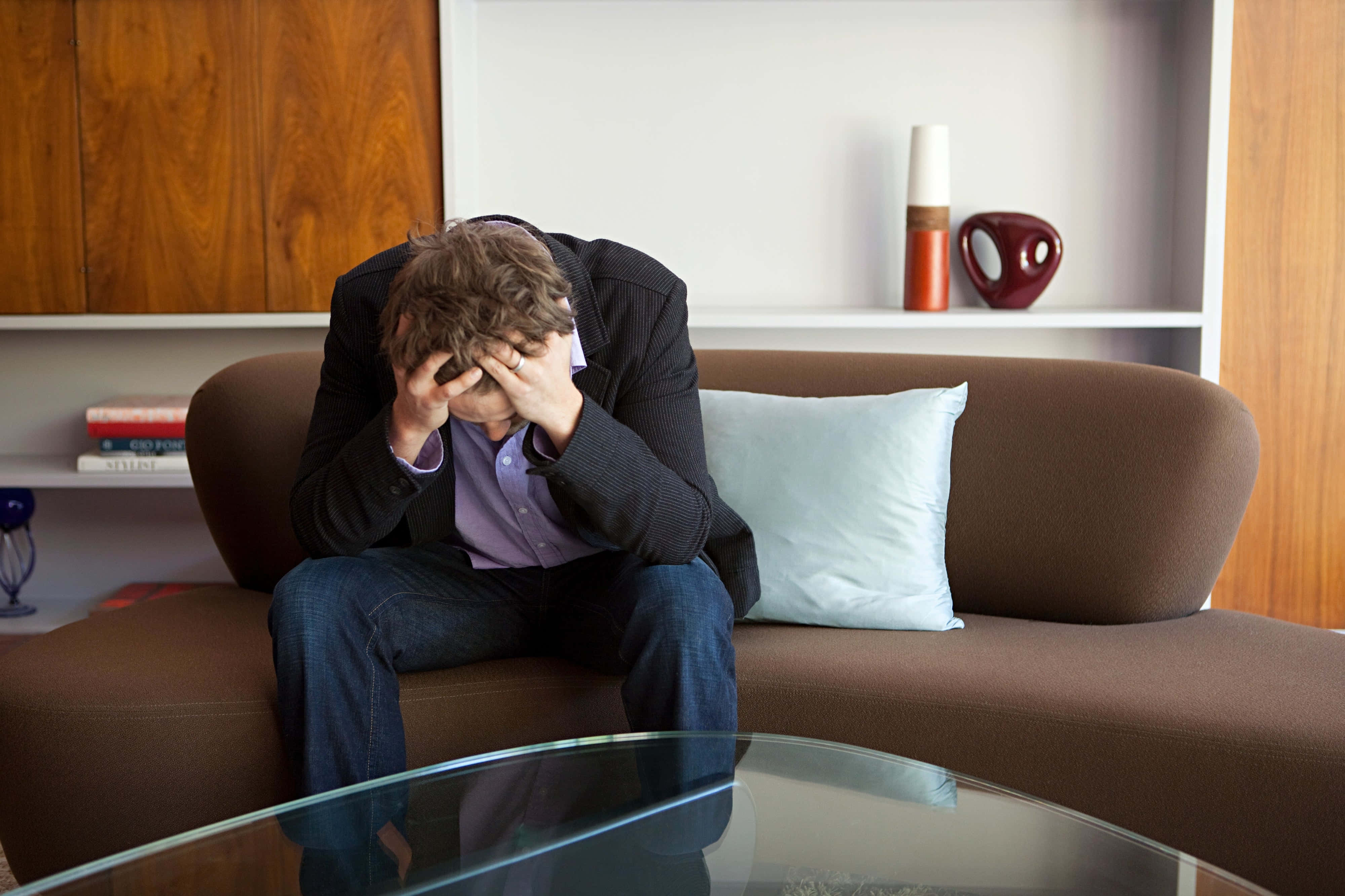 Depressed 桌布 壁紙 — 一名身穿西裝的男子癱坐在棕色沙發上，雙手掩面，神情痛苦，身處一個有木架和玻璃咖啡桌的現代客廳。
