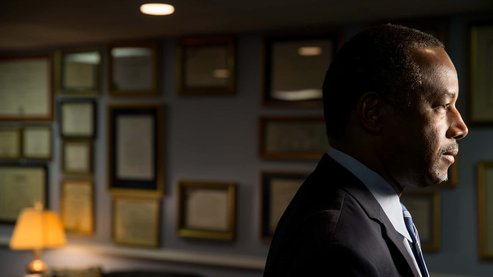 美國 桌布 壁紙 — 一名身穿西裝的男子站在光線昏暗的辦公室內，牆上掛著裱框證書，附近有一盞溫暖的燈在發光。