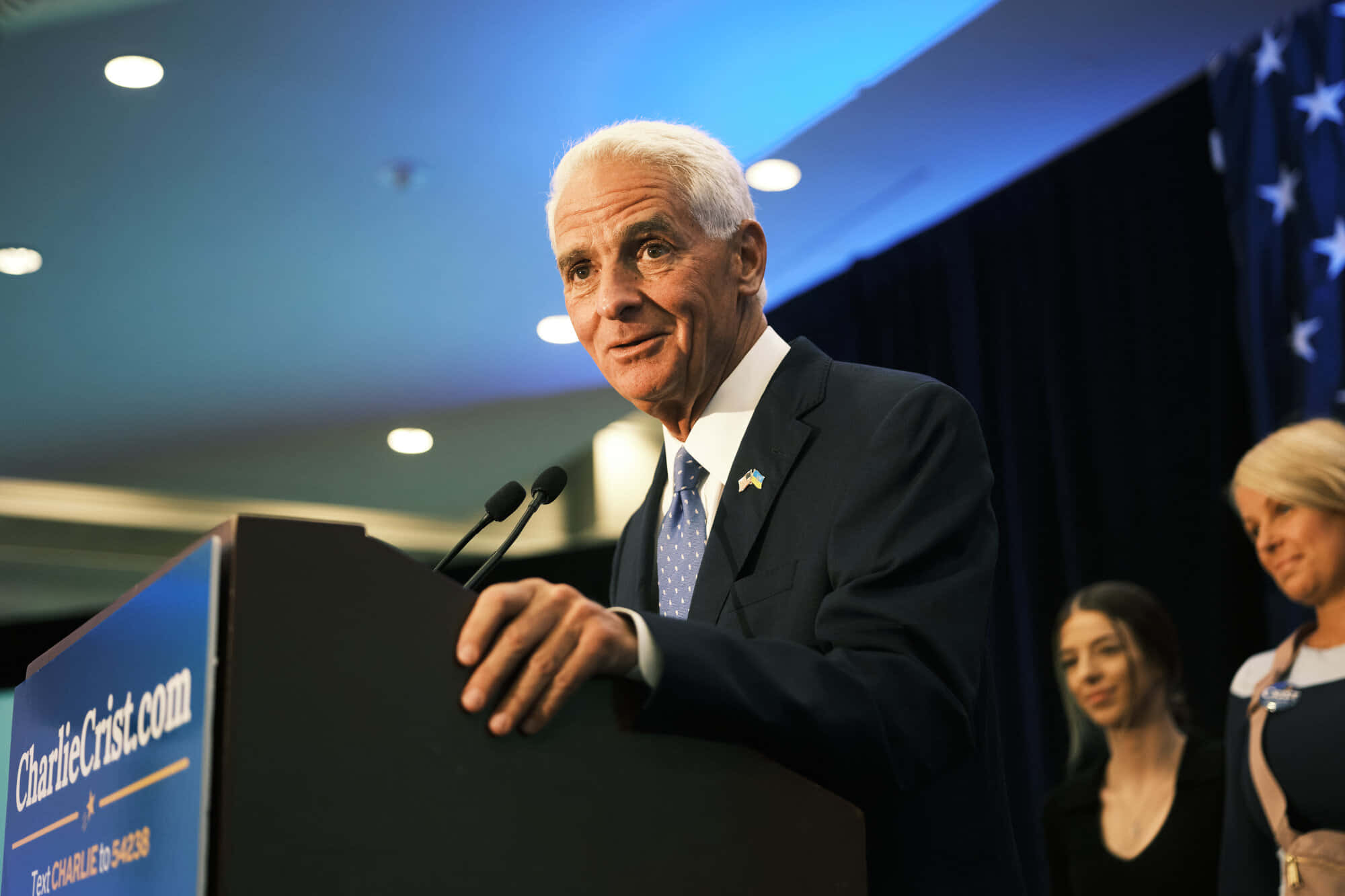 桌布 壁紙 — 一名身穿西裝的男子站在設有「CharlieCrist.com」標誌的講台上，在藍色舞台燈光下向觀眾發言。