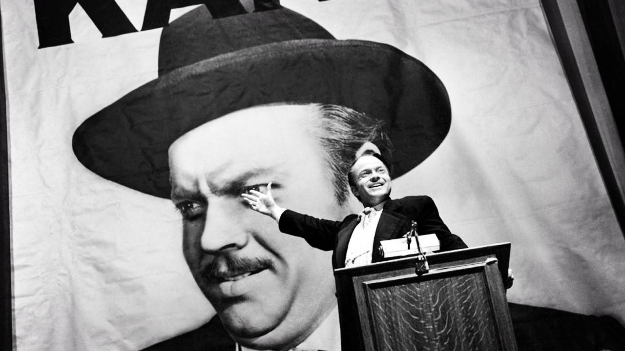 Citizen Kane 桌布 壁紙 — 一名身穿西裝的男子站在講台上，向身後自己巨大的黑白肖像做手勢。