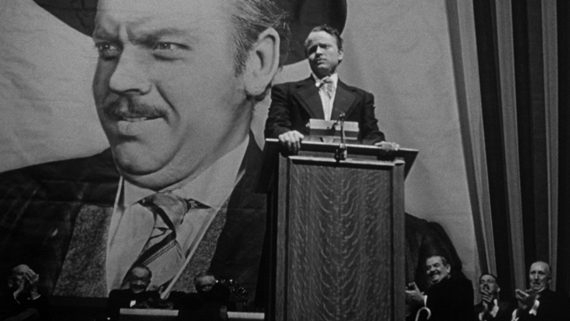 Citizen Kane 桌布 壁紙 — 一名身穿西裝的男子站在講台上，面前是一幅巨大的鬍鬚男子肖像，場景戲劇性且黑白分明。