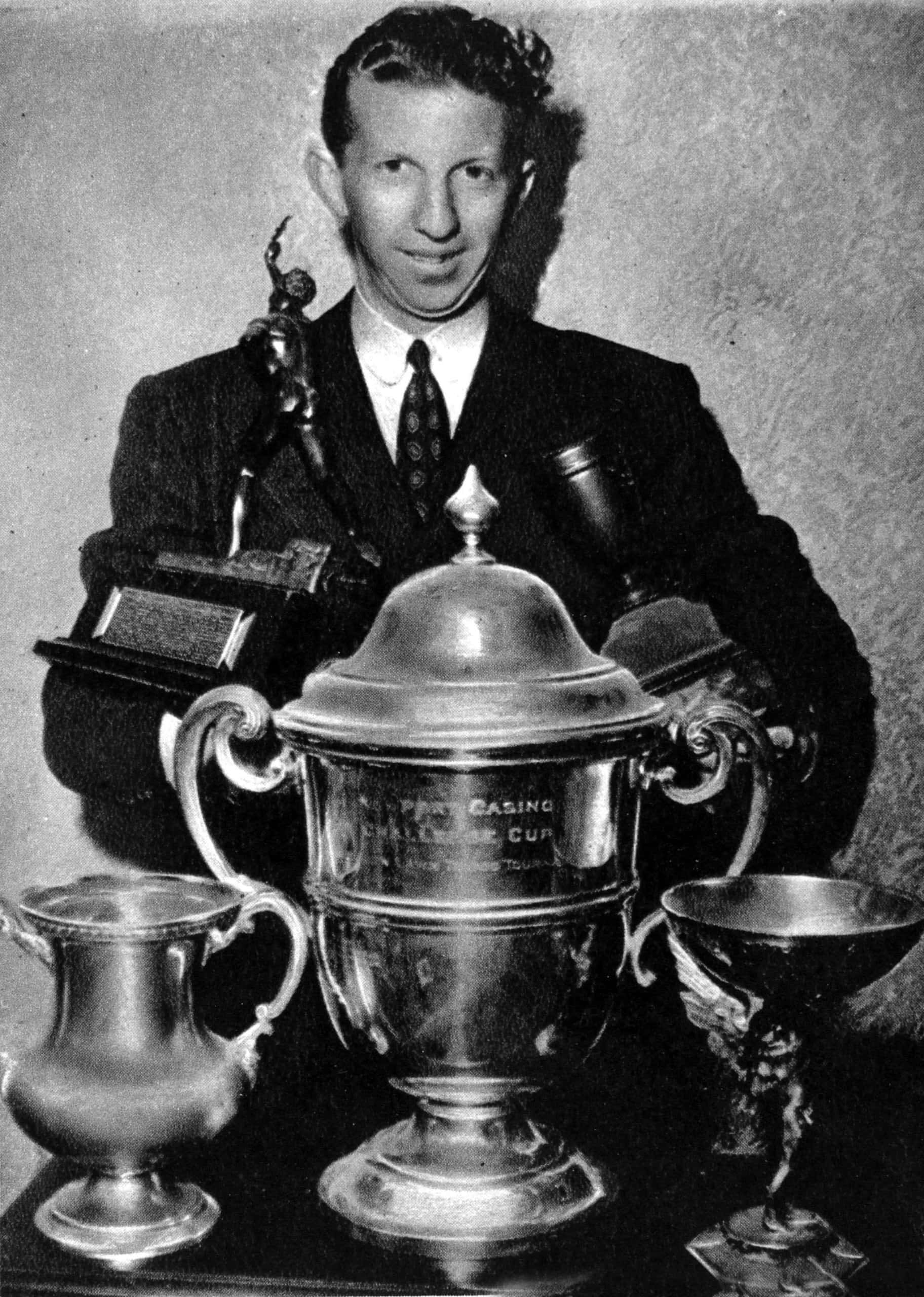 Trophy 桌布 壁紙 — 一名身穿西裝的男子自豪地展示多個銀色獎盃，包括桌上一個有蓋的大獎盃。