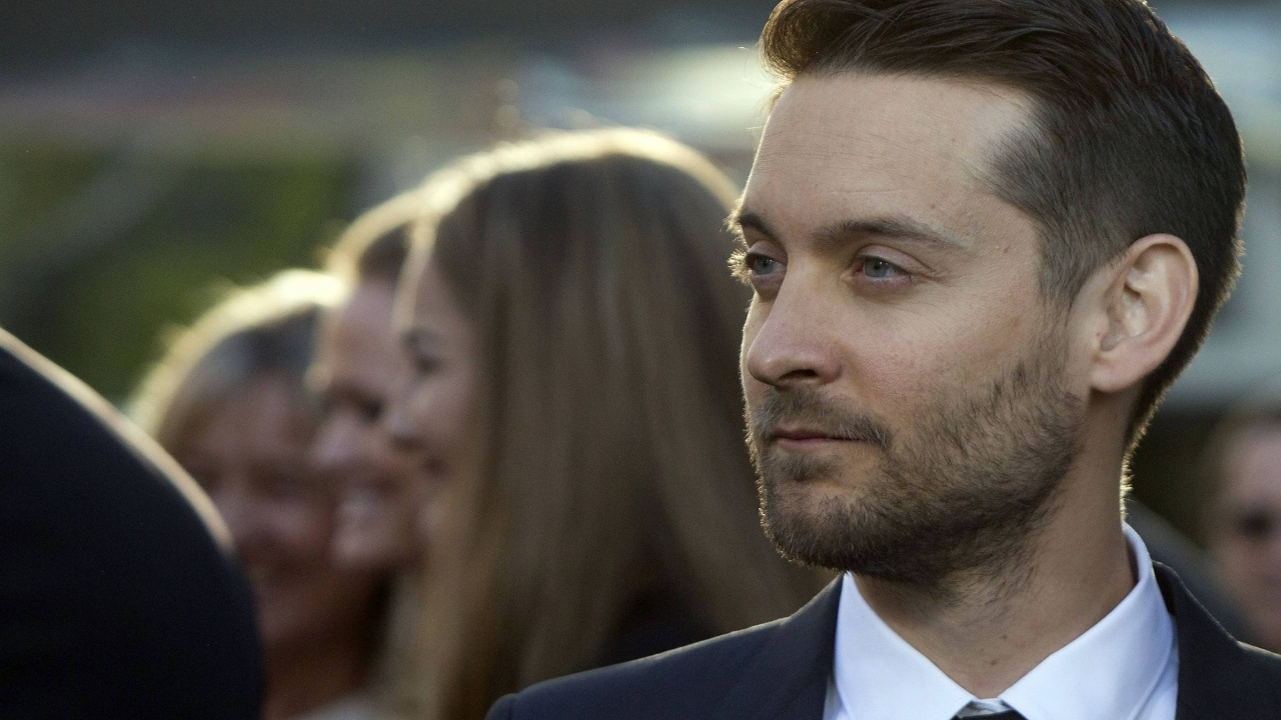 Tobey Maguire 桌布 壁紙 — 一名身穿西裝的男子若有所思地望向一旁，模糊的人們在他身後柔和的金色光芒中微笑。