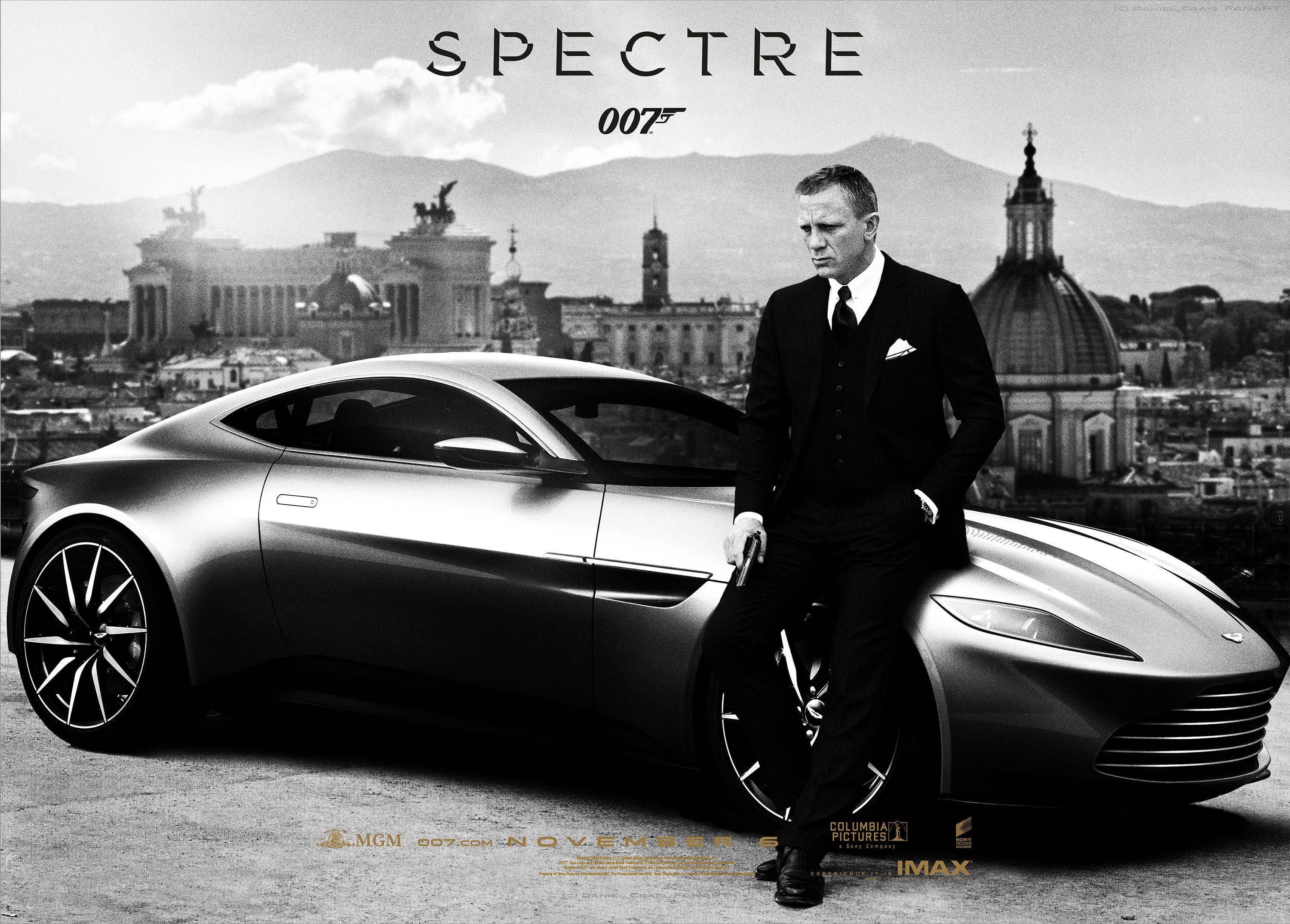 Daniel Craig 桌布 壁紙 — 一名身穿西裝的男子靠在一輛流線型的銀色跑車上，背景是城市天際線和山脈。