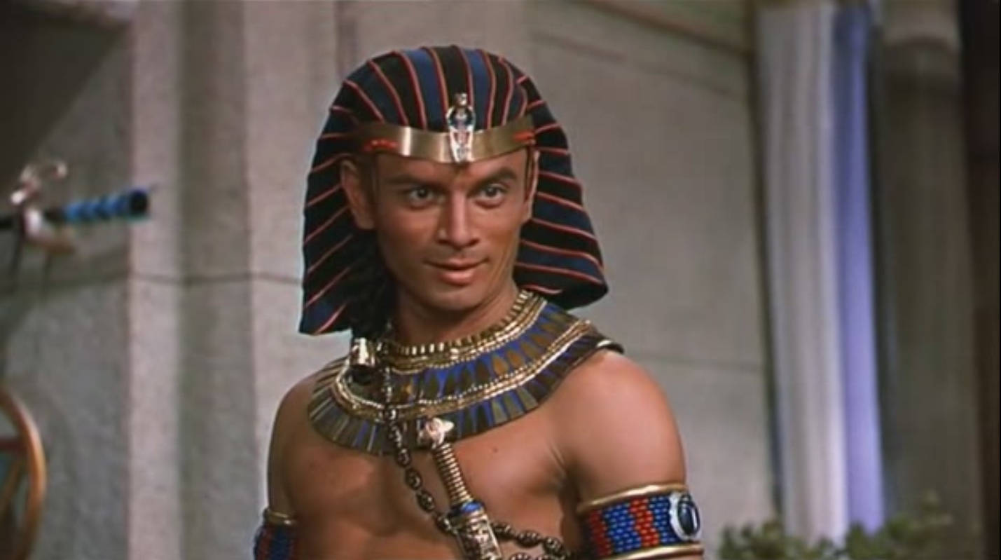 egyptian 桌布 壁紙 — 一名身穿詳細古埃及皇室服飾、戴著條紋頭飾和金飾的男子，站在石牆環境中。