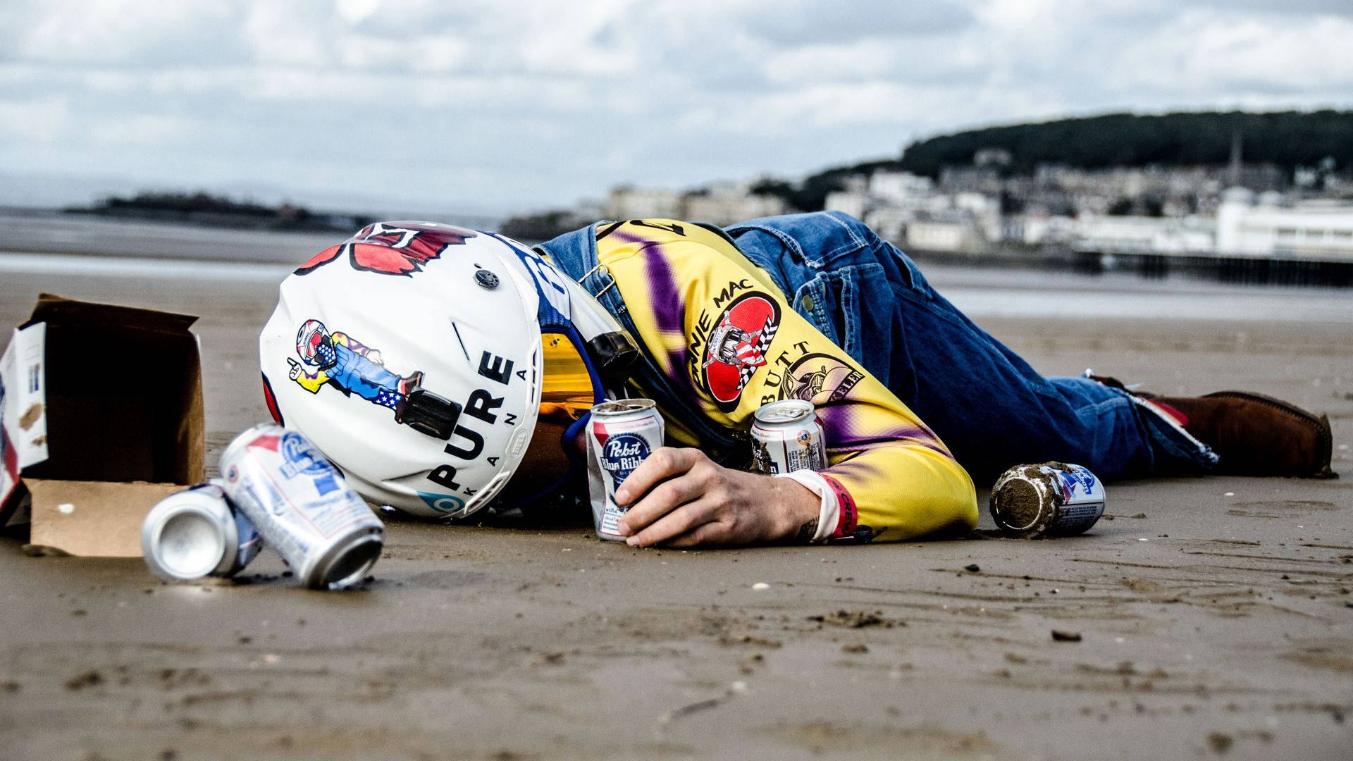 Ronnie Mac 桌布 壁紙 — 一名身穿賽車裝備的人躺在濕漉漉的海灘上不省人事，周圍散落著空啤酒罐和一個紙箱。