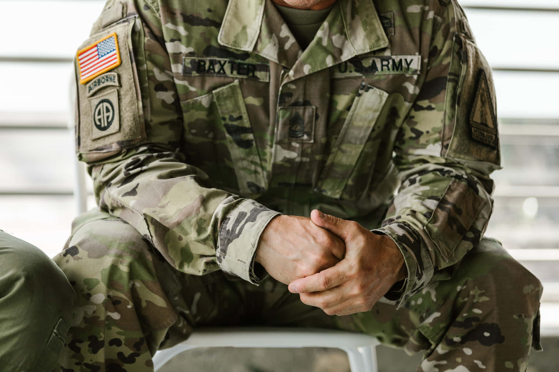 壁紙 — 一名身穿迷彩制服的美國陸軍士兵雙手合十坐在白色椅子上，名牌和空降徽章清晰可見。