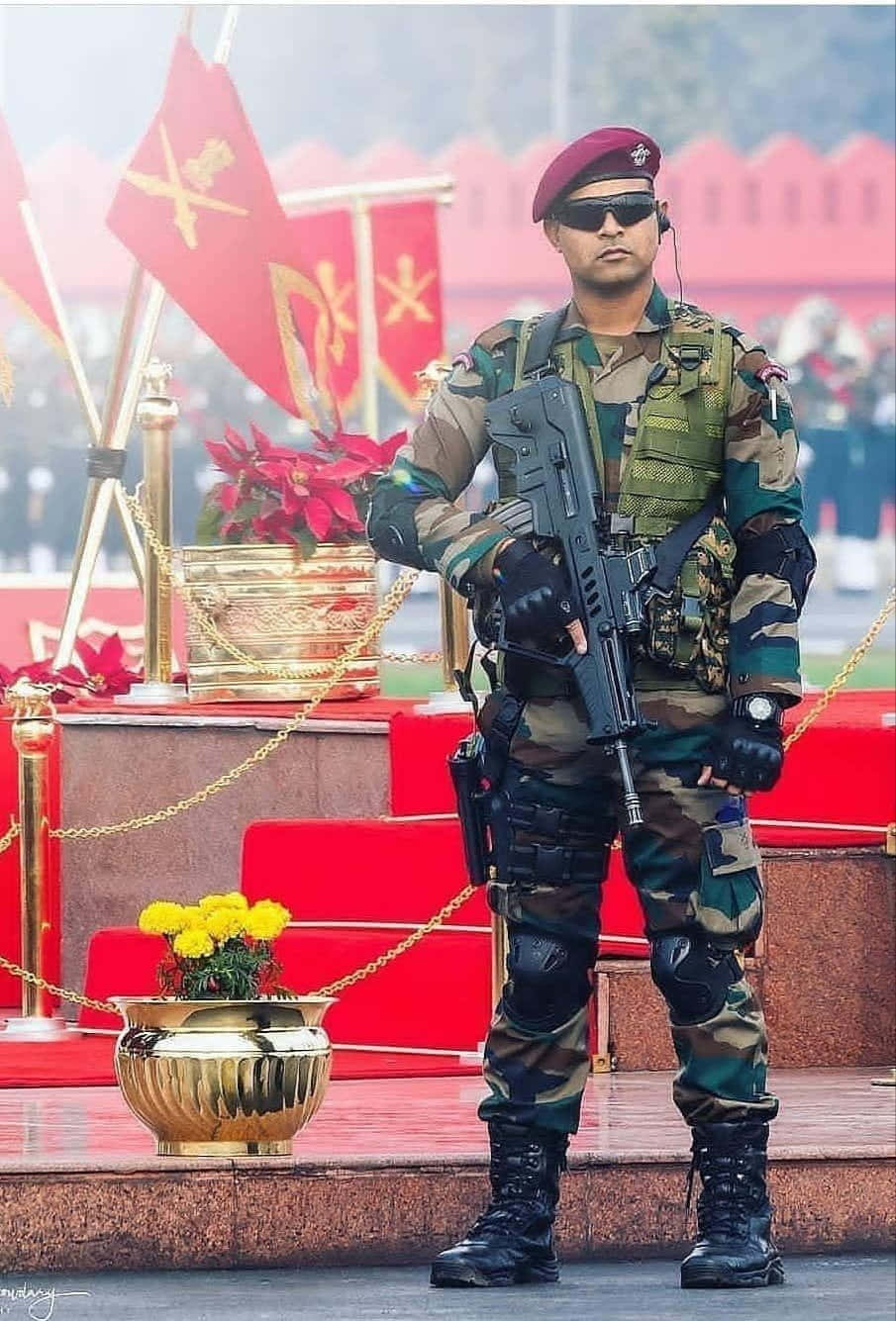 印度軍隊 桌布 壁紙 — 一名身穿迷彩服和紅色貝雷帽的士兵，在帶有金色花盆和旗幟的紅色台階旁持步槍立正站立。