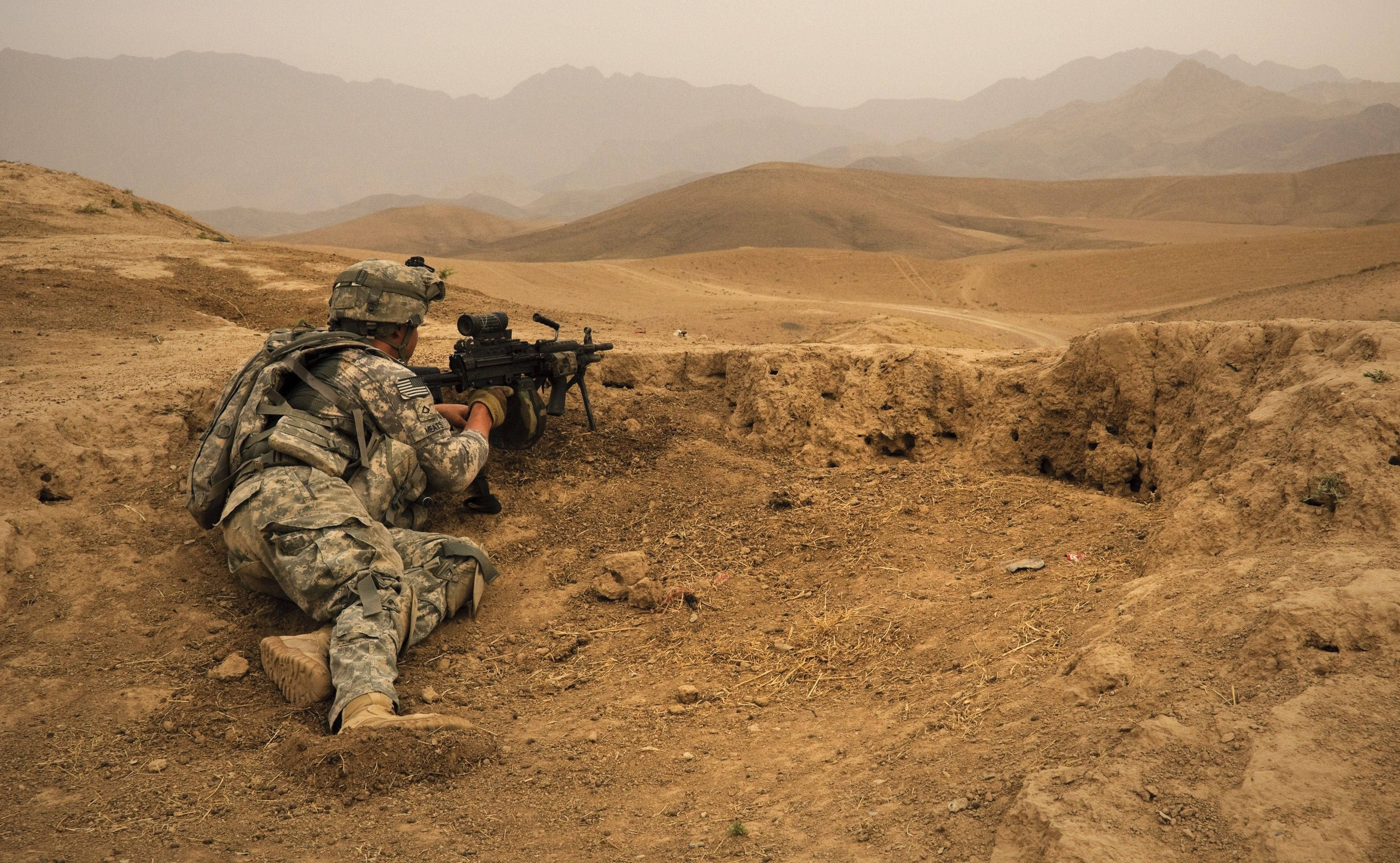 US 桌布 壁紙 — 一名身穿迷彩服的士兵俯臥在塵土飛揚的戰壕中，瞄準機槍，越過一片荒涼的沙漠景觀，遠處有朦朧的山脈。