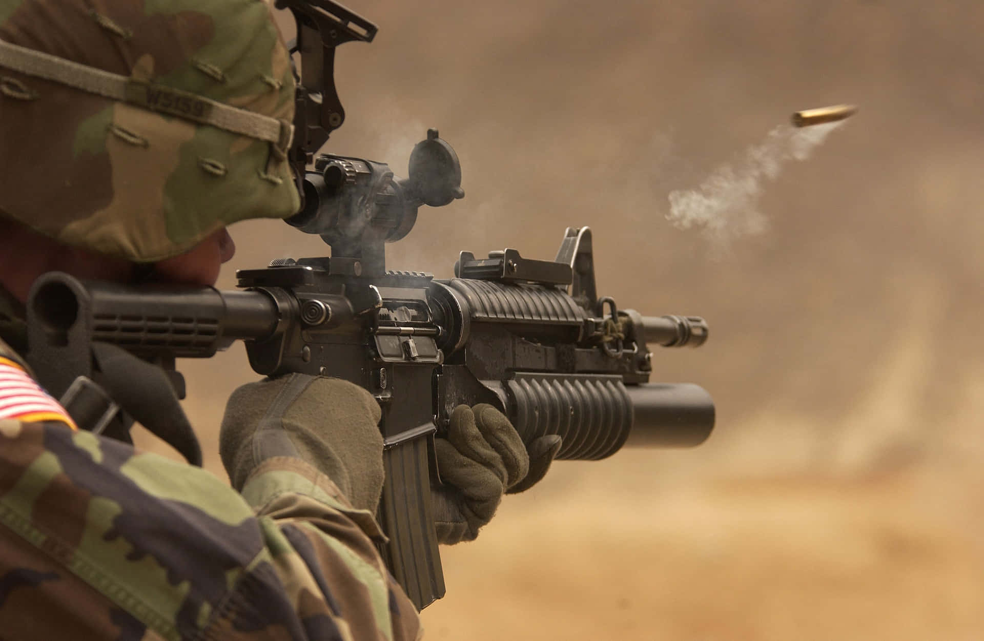  壁紙 — 一名身穿迷彩服的士兵在塵土飛揚的沙漠中發射突擊步槍，子彈留下煙霧軌跡。