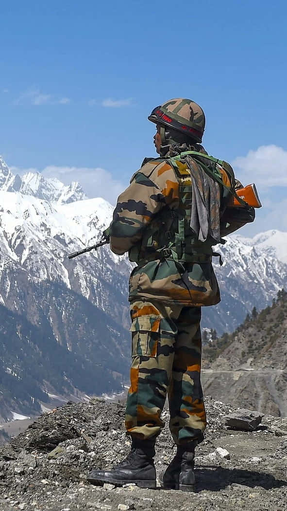 印度軍隊 桌布 壁紙 — 一名身穿迷彩服的士兵在岩石山脊上站崗，俯瞰著晴朗藍天下白雪覆蓋的山峰。