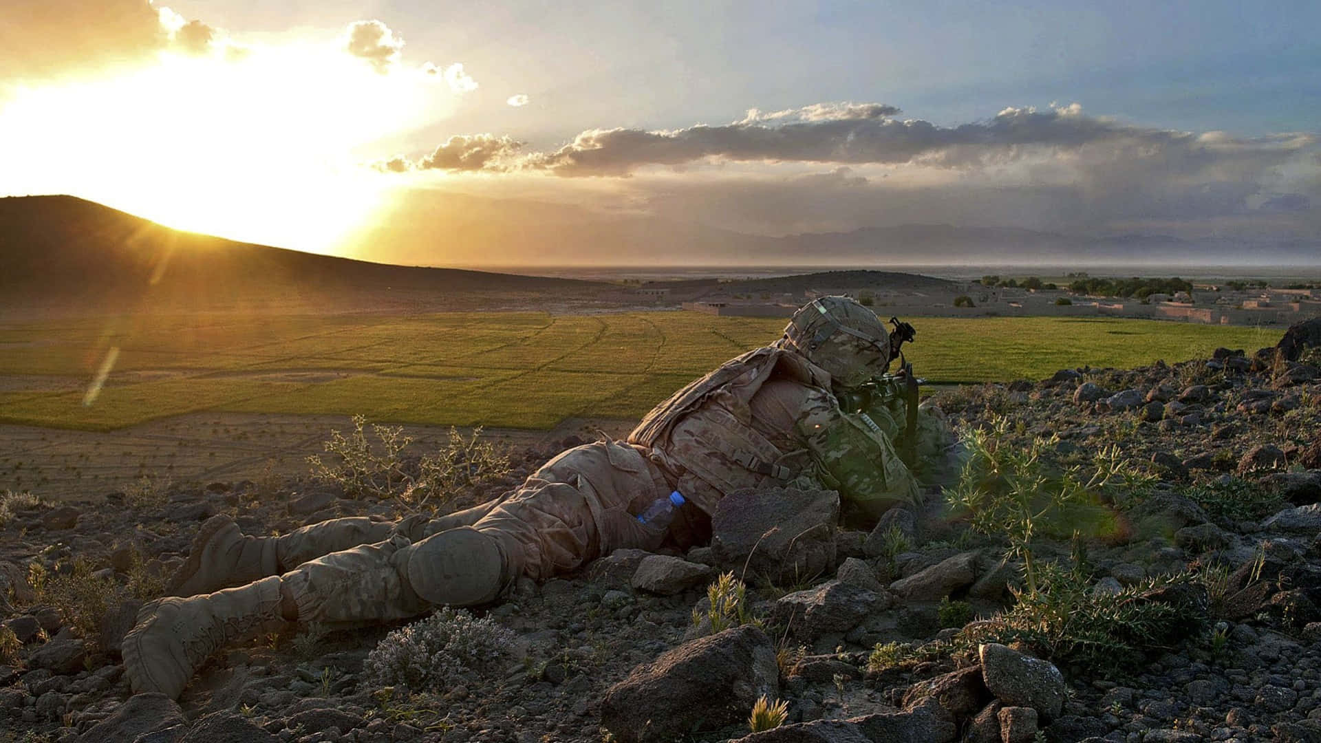 Afghanistan 桌布 壁紙 — 一名身穿迷彩服的士兵在日落時分俯臥在岩石地形上，俯瞰著金色天空下廣闊的綠色田野和遠處的山脈。