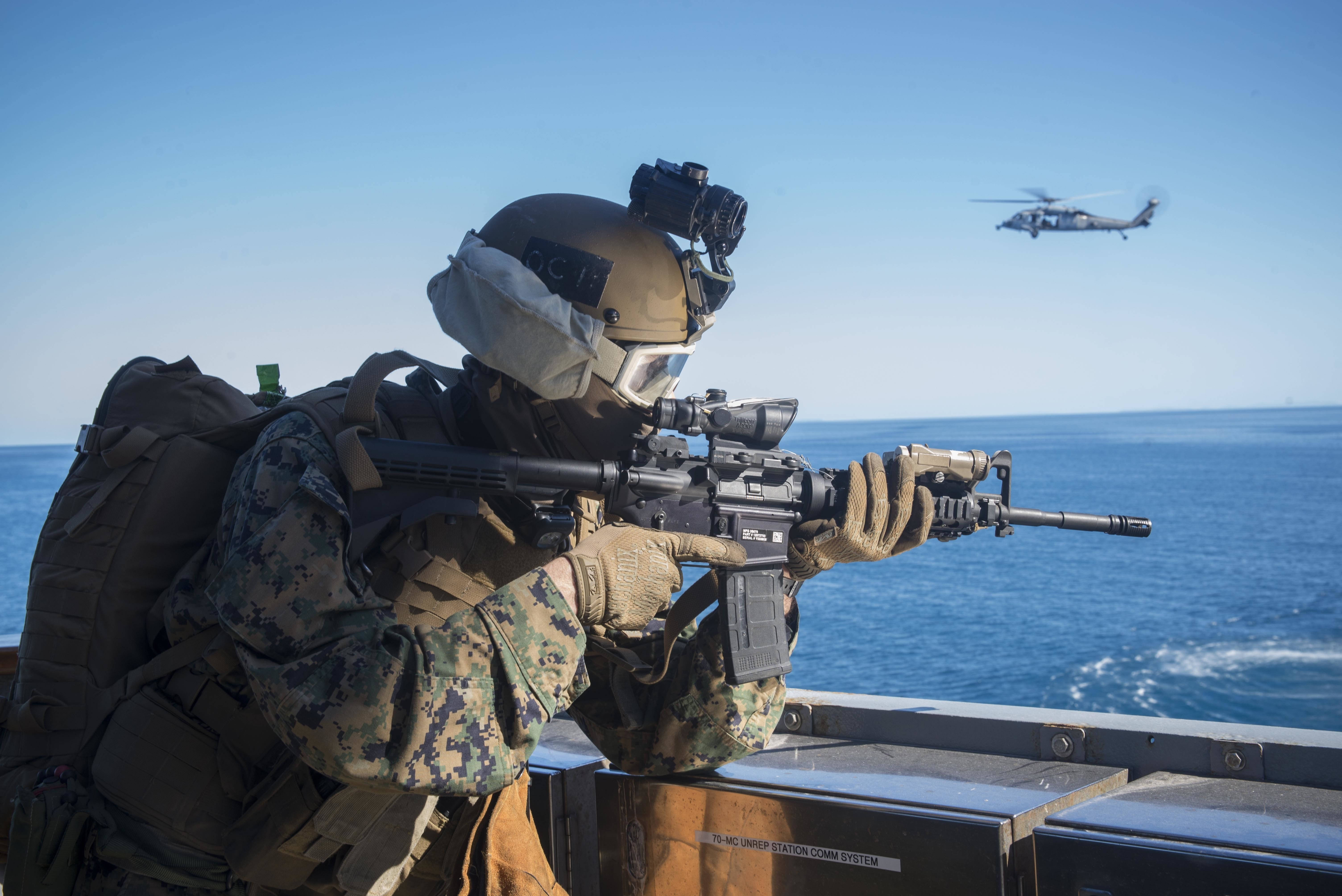 Marine 桌布 壁紙 — 一名身穿迷彩服的士兵在船甲板上瞄準步槍，一架直升機在清澈藍天下的海洋上空飛行。