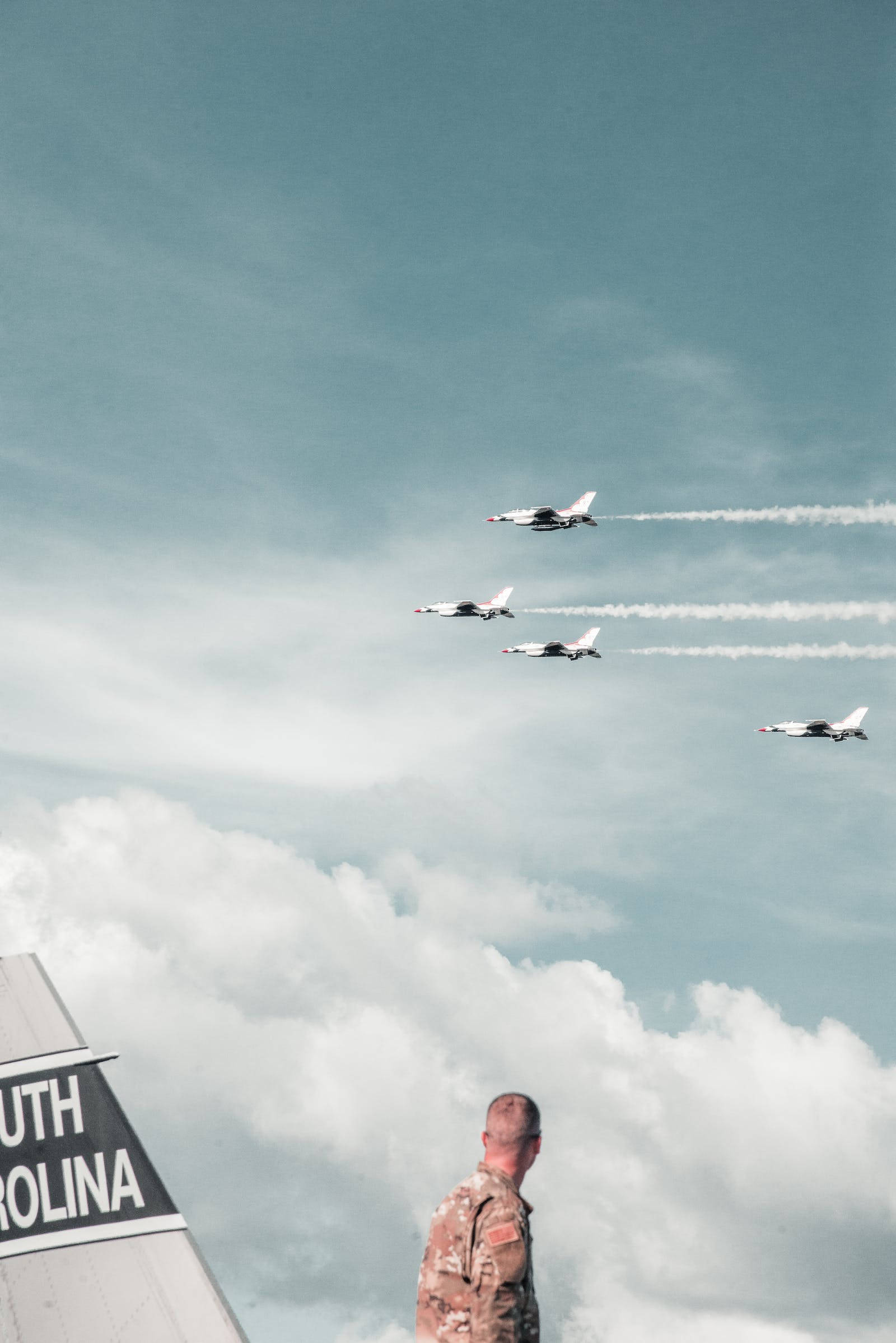 Air Force 桌布 壁紙 — 一名身穿迷彩服的士兵站立觀看四架戰鬥機在頭頂編隊飛行，在陰雲密佈的天空中留下白色煙跡。