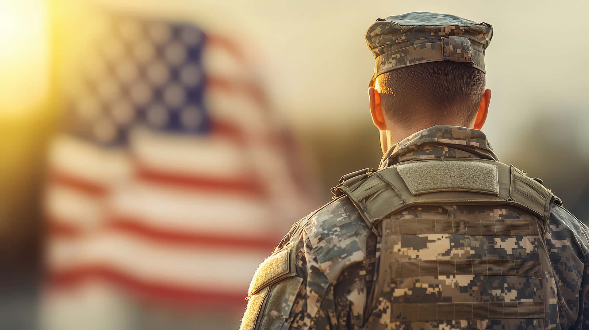A military soldier in camouflage uniform stands with his back turned, looking at the American flag 壁紙 — 一名身穿迷彩服的士兵背對鏡頭，面向日落時模糊的美國國旗。