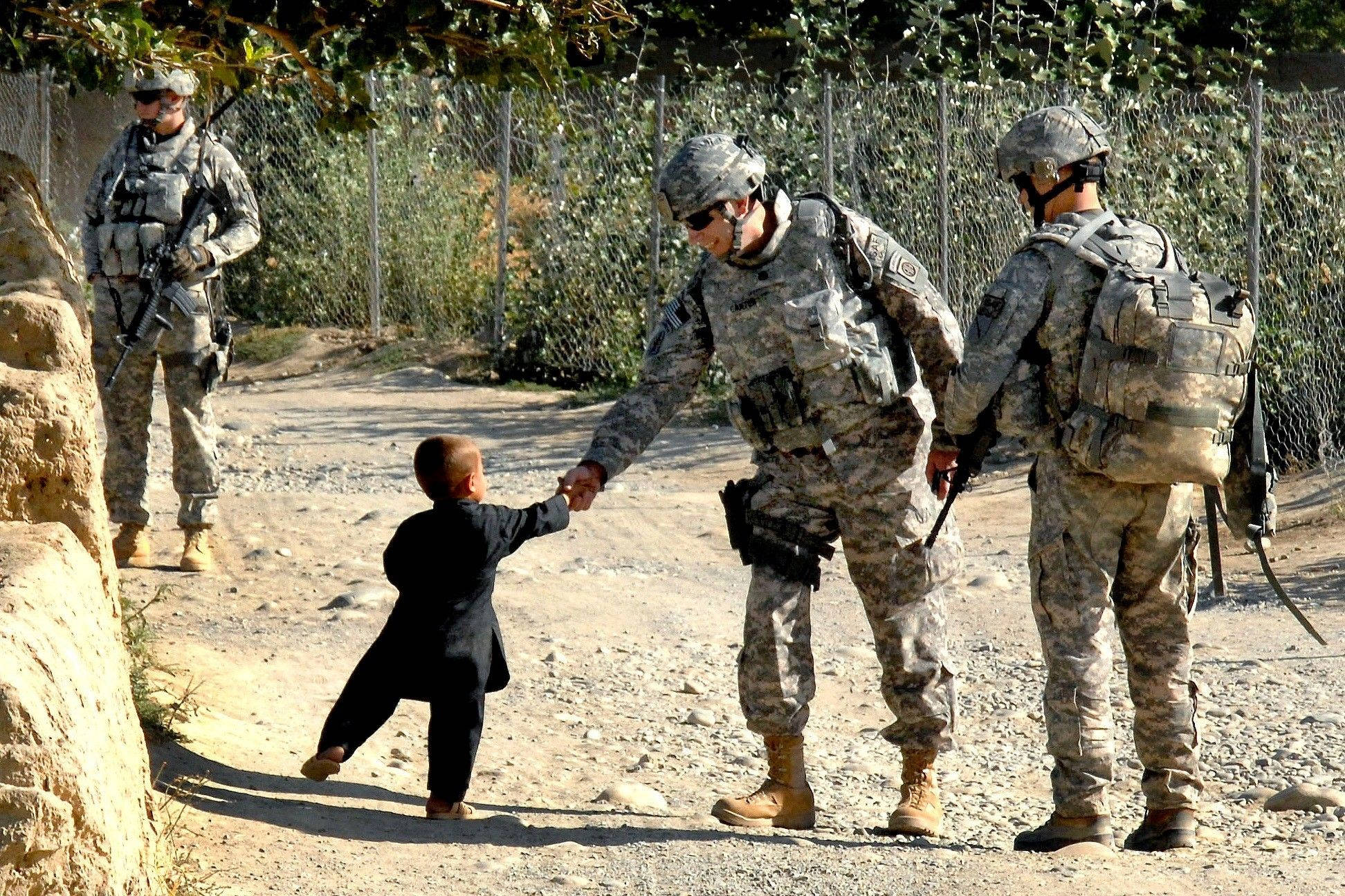 US 桌布 壁紙 — 一名身穿迷彩服的士兵跪在塵土飛揚的小路上，與一個身穿黑色西裝的小孩握手，旁邊有圍欄。