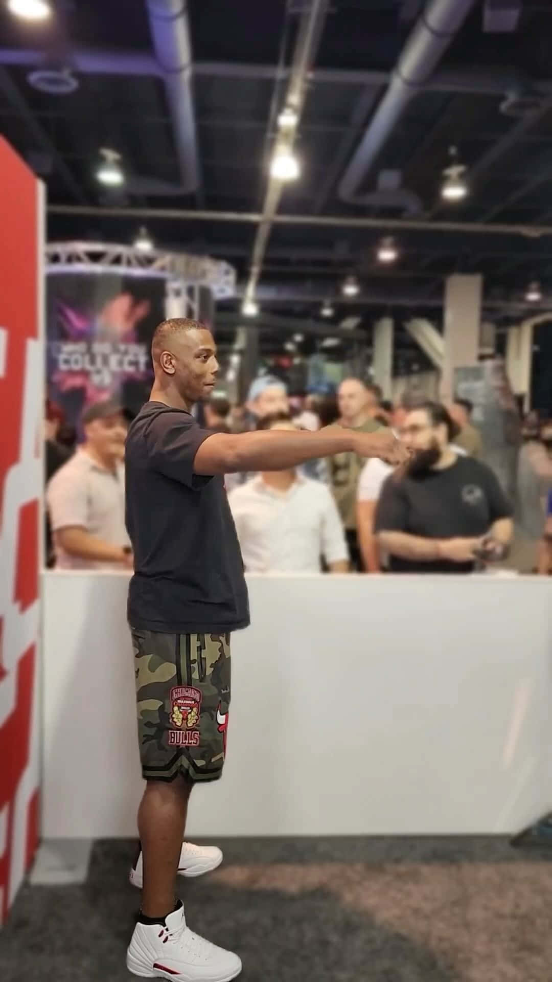 MMA 桌布 壁紙 — 一名身穿迷彩短褲和白色運動鞋的男子，在擠滿人的會議廳中擺姿勢，頭頂有外露的管道和明亮的燈光。