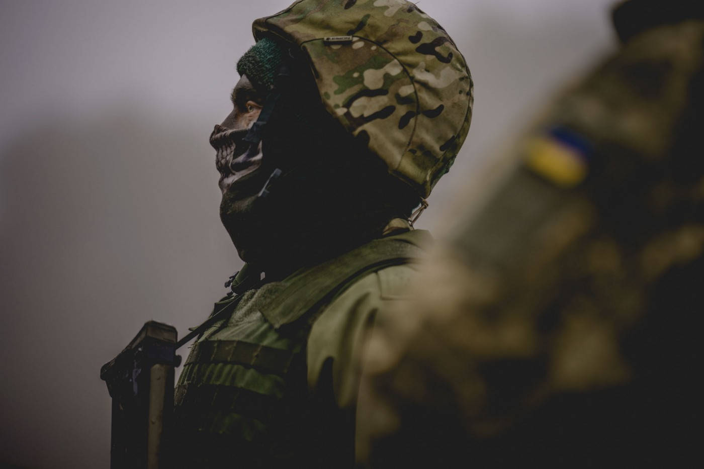 Ukraine 桌布 壁紙 — 一名身穿迷彩裝備和骷髏圖案面罩的士兵，在柔和的灰色背景前側面站立。