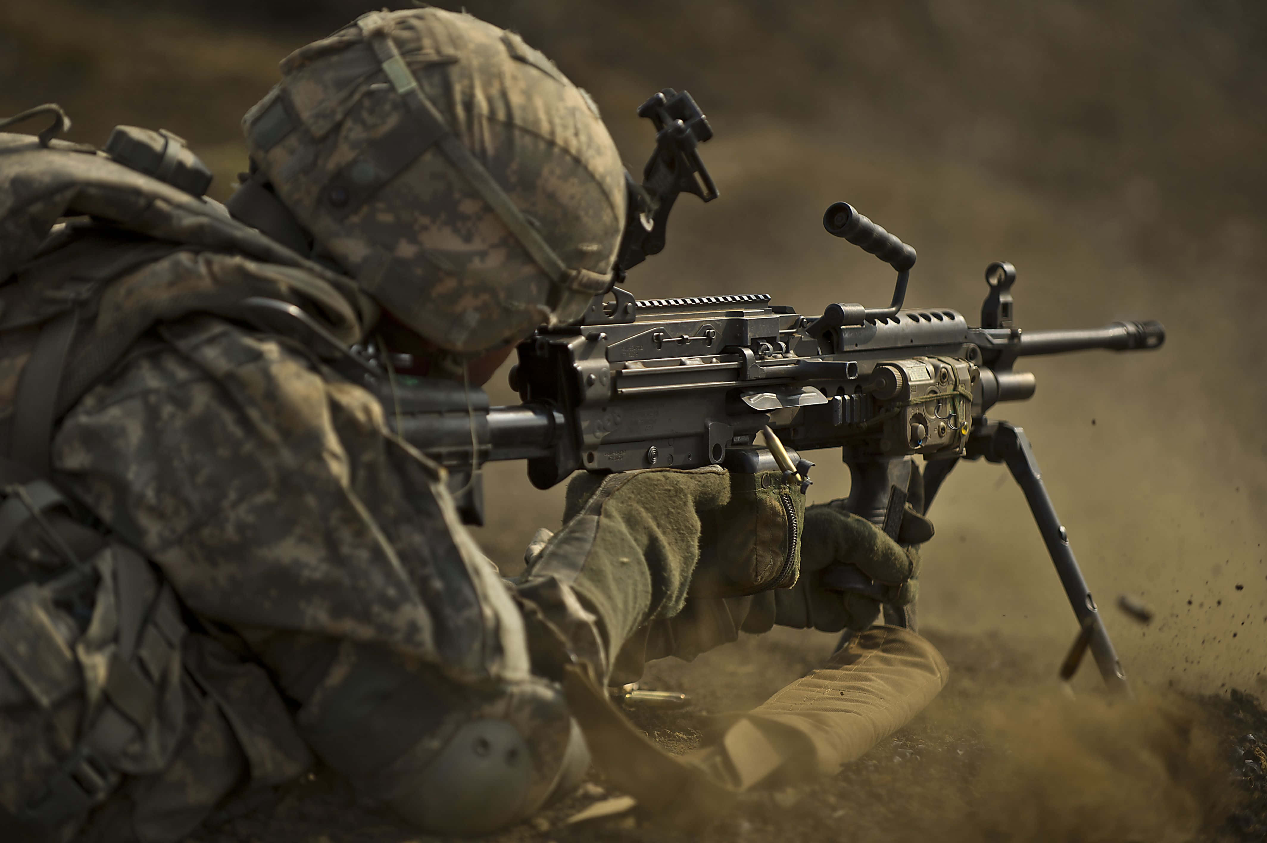 軍隊 桌布 壁紙 — 一名身穿迷彩裝備的士兵以俯臥姿勢發射重型機槍，在荒蕪的田野中揚起灰塵。
