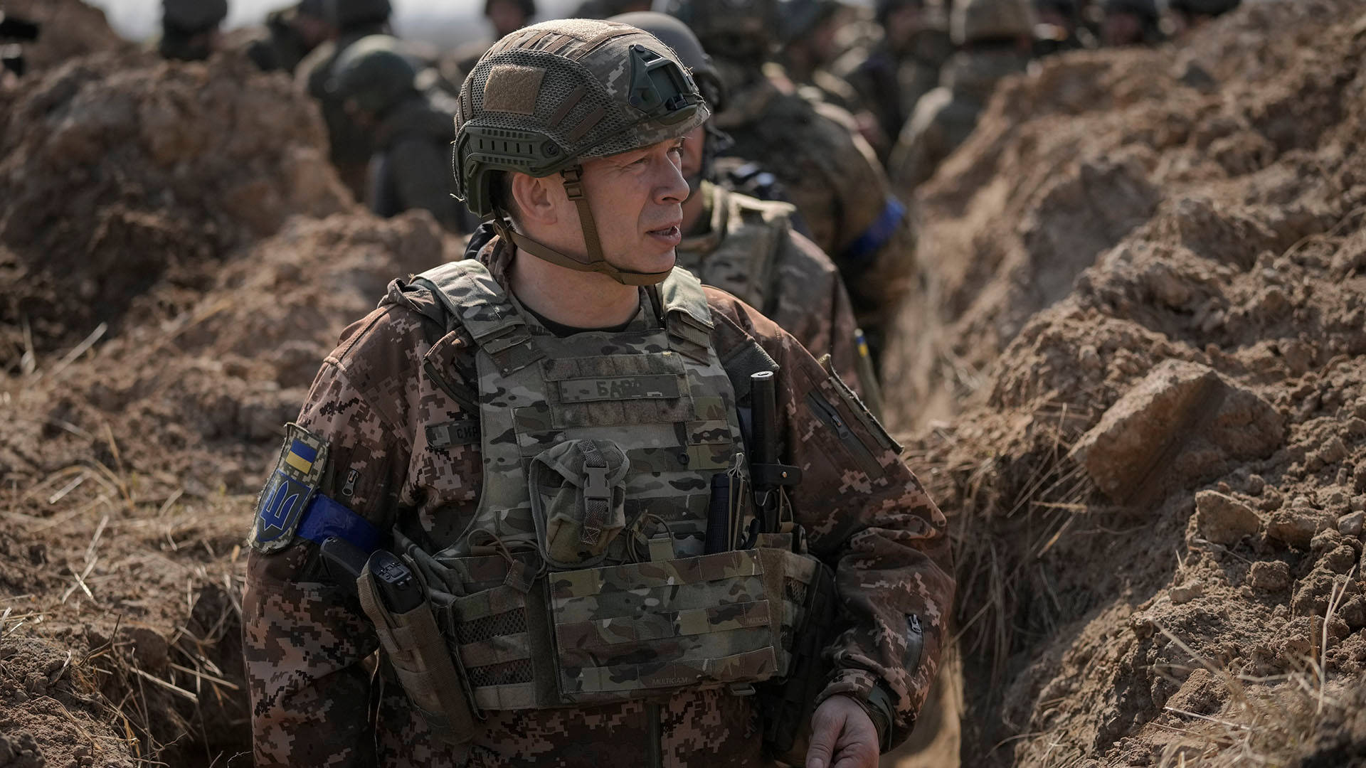 軍隊 桌布 壁紙 — 一名身穿迷彩裝備的士兵在泥濘的戰壕中保持警覺，背景有其他部隊。