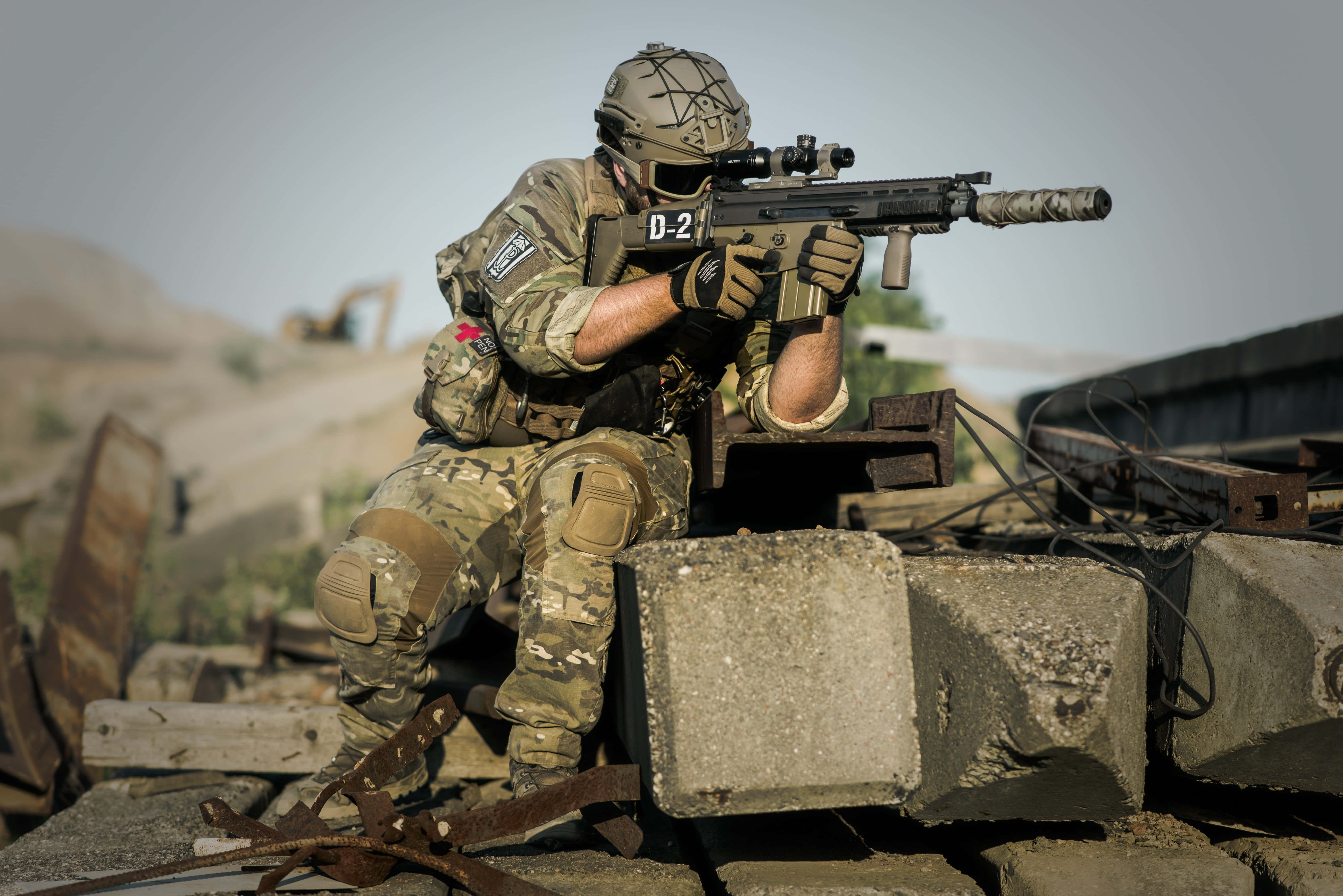 soldier 桌布 壁紙 — 一名身穿迷彩裝備的士兵在陽光照耀的戰區中，從混凝土瓦礫後方瞄準步槍。