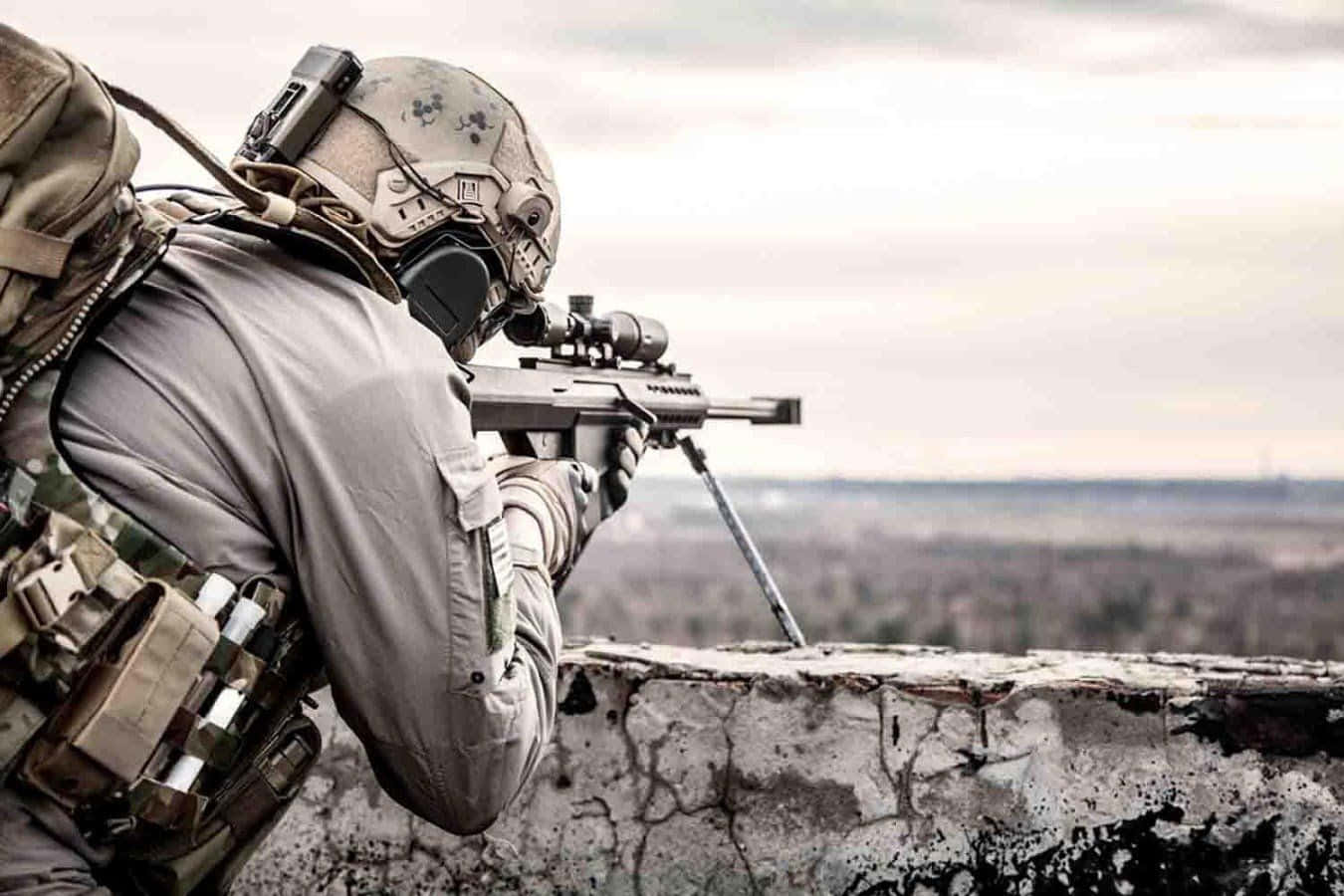 Tactical 桌布 壁紙 — 一名身穿迷彩裝備的士兵從風化的石牆上瞄準狙擊步槍，俯瞰陰天下一片廣闊的開闊地帶。