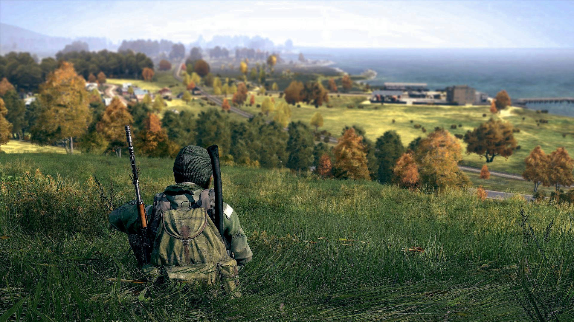 DayZ 桌布 壁紙 — 一名身穿迷彩裝備的士兵蹲在草坡上，俯瞰著一個有秋樹和遠洋的沿海村莊。