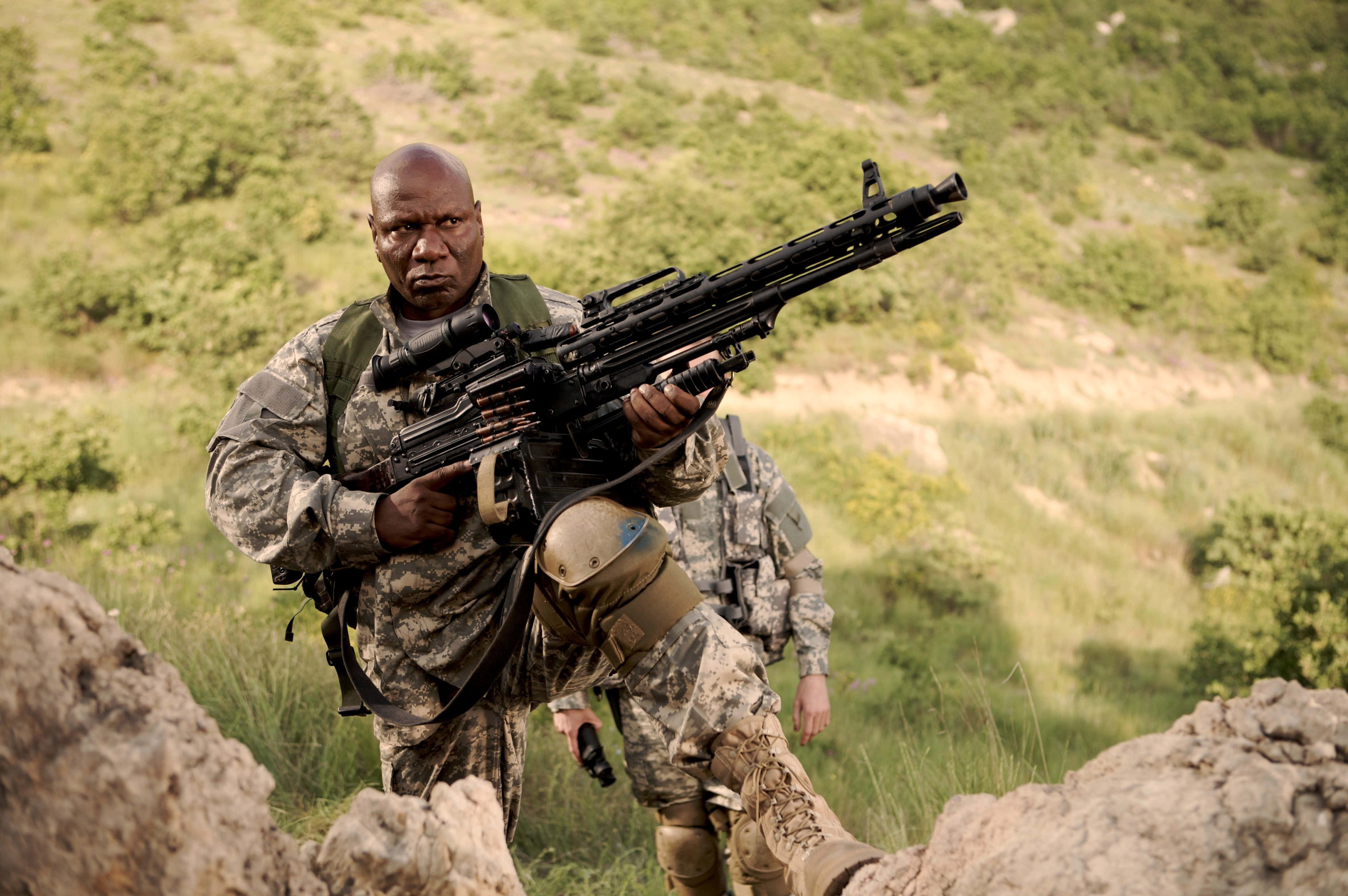 Ving Rhames 桌布 壁紙 — 一名身穿迷彩裝備的士兵蹲在陽光照耀的草坡岩石後，手持重型機槍。
